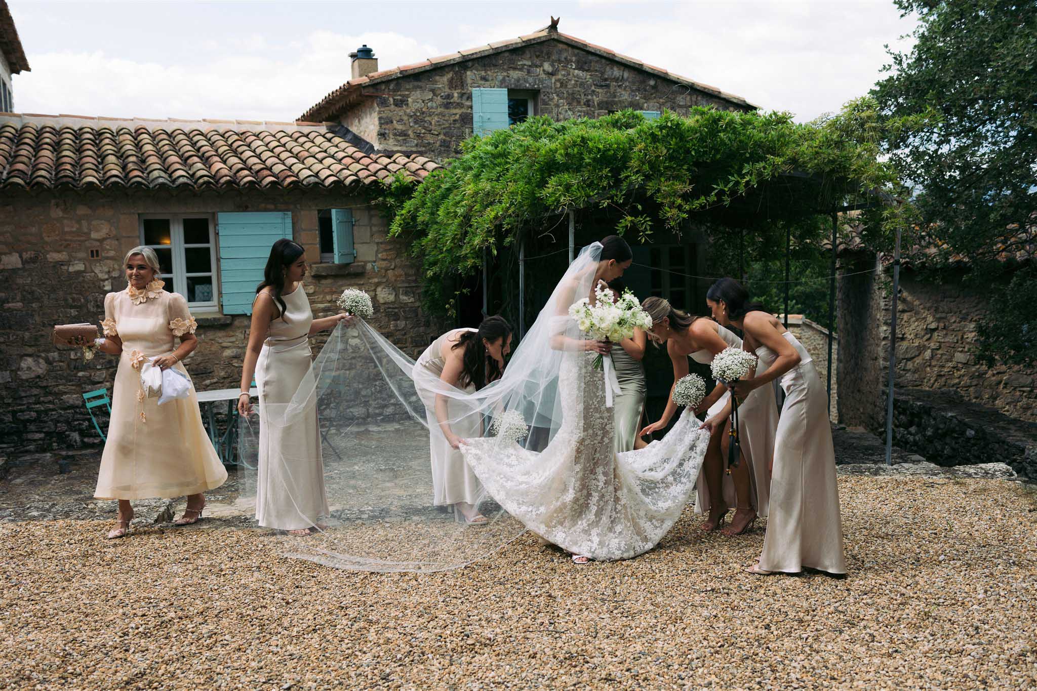 Bride in white lace gown with five bridesmaids in champagne dresses arranging cathedral train in stone courtyard