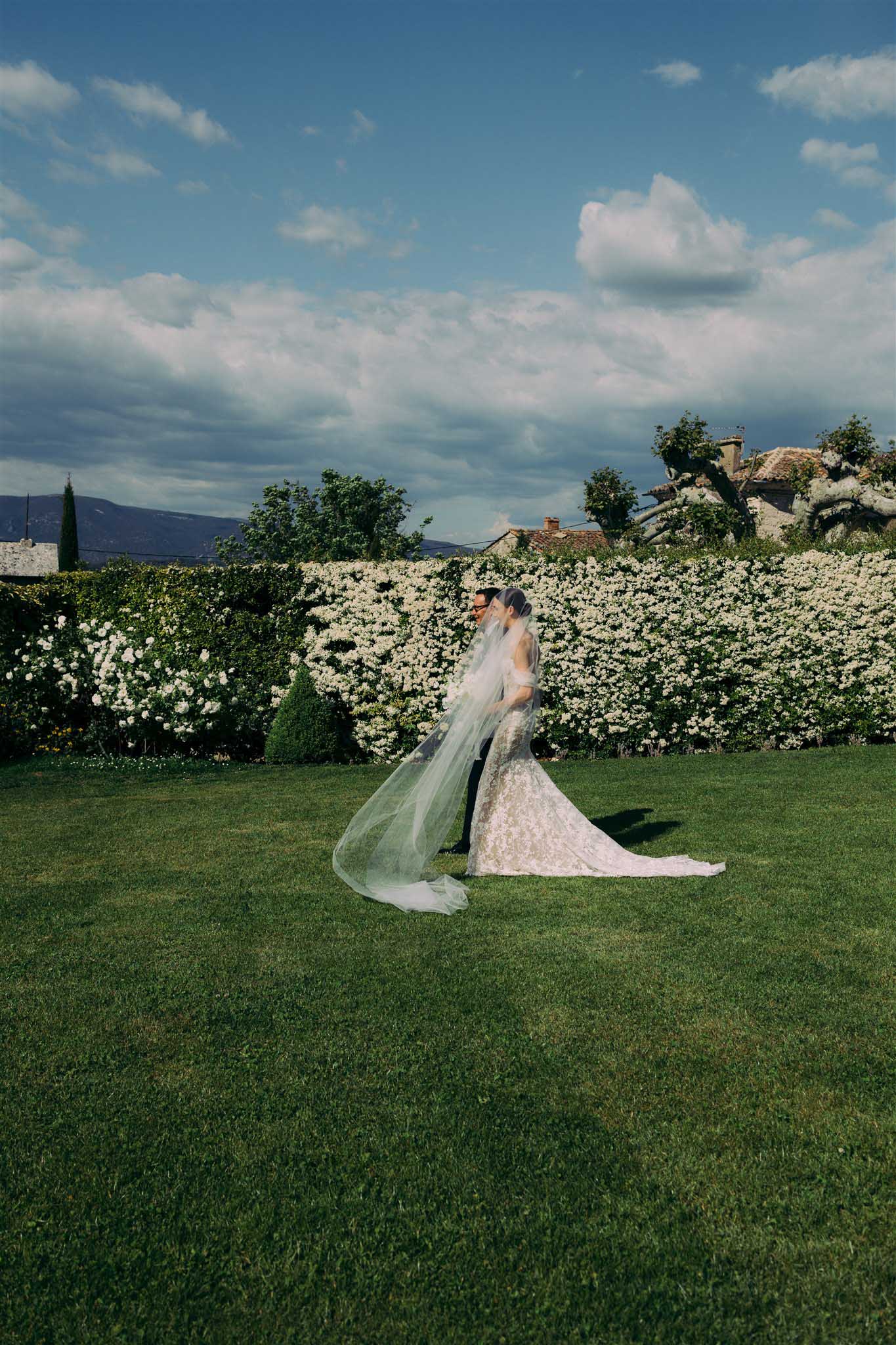 Bride in ivory lace dress with sage green veil stands before a white flowering hedge at a countryside villa