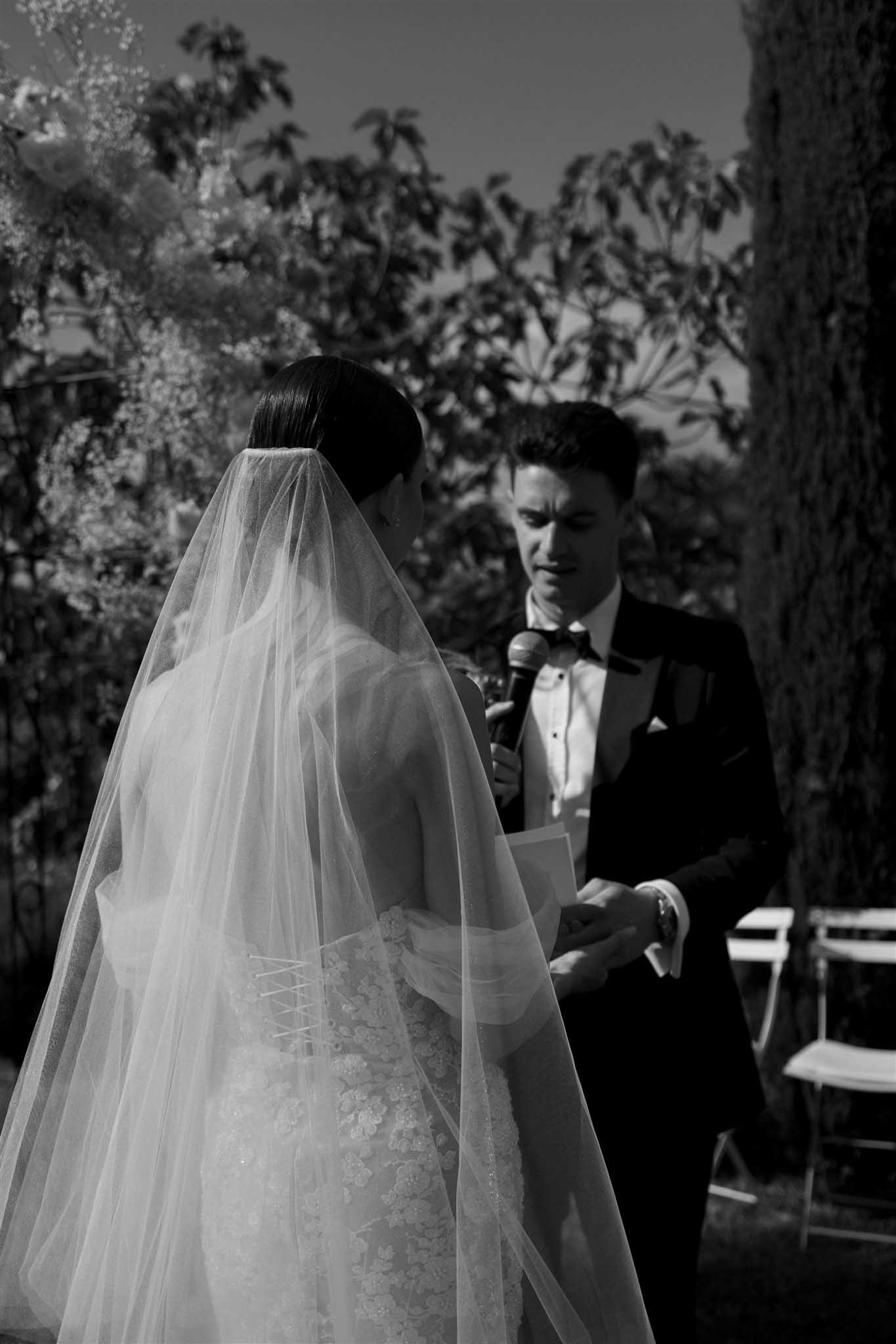 Black and white outdoor ceremony, groom reading vows near cypress trees as bride in embellished bodice listens