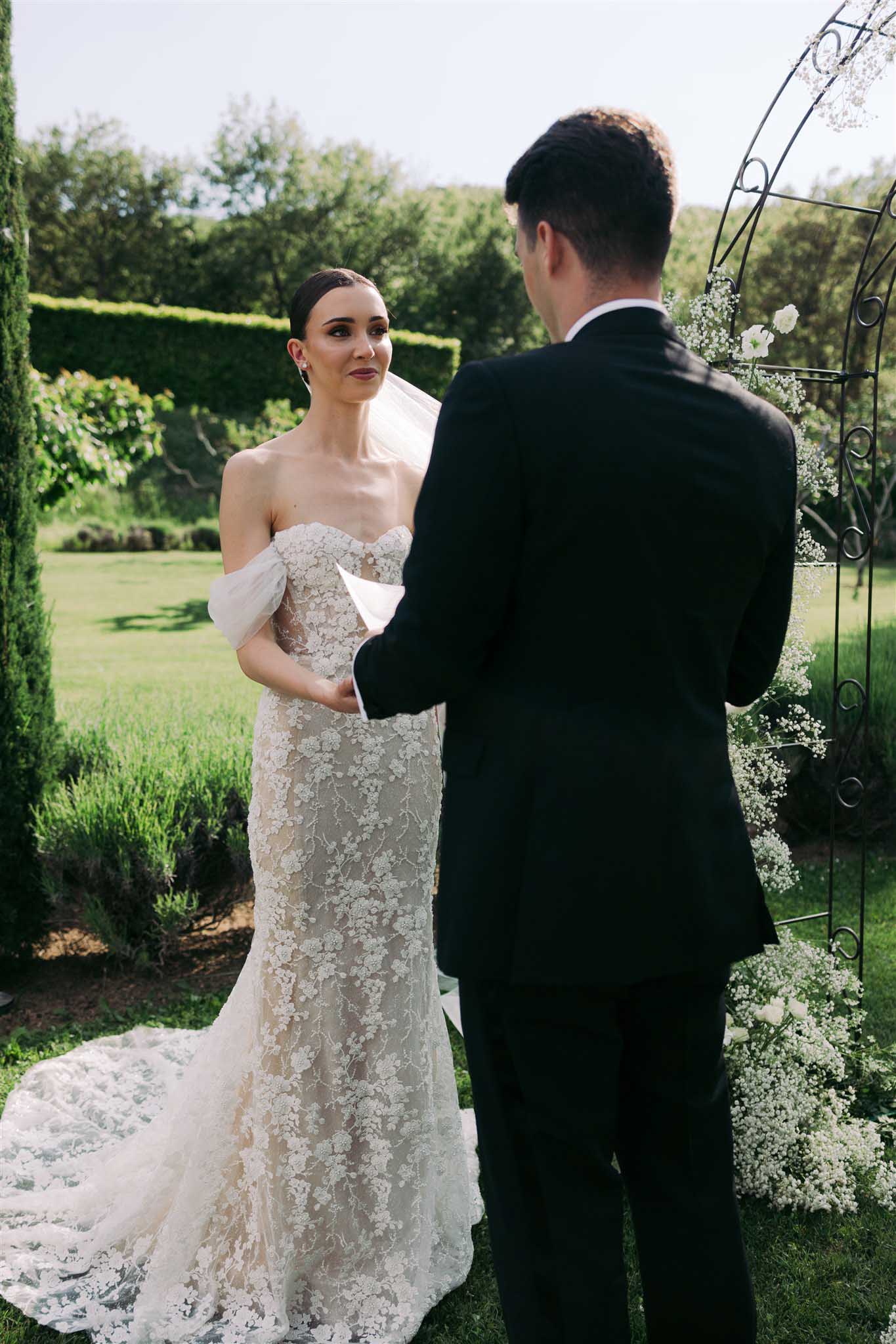 Bride and groom exchanging vows beneath wrought-iron arch with baby's breath in formal garden