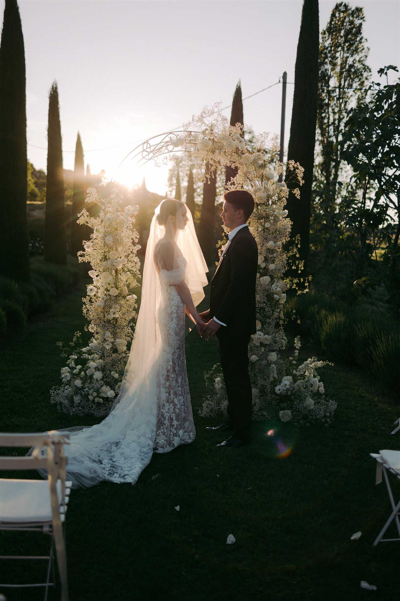 Bride and groom beneath baby's breath floral arch at golden sunset ceremony with cypress trees