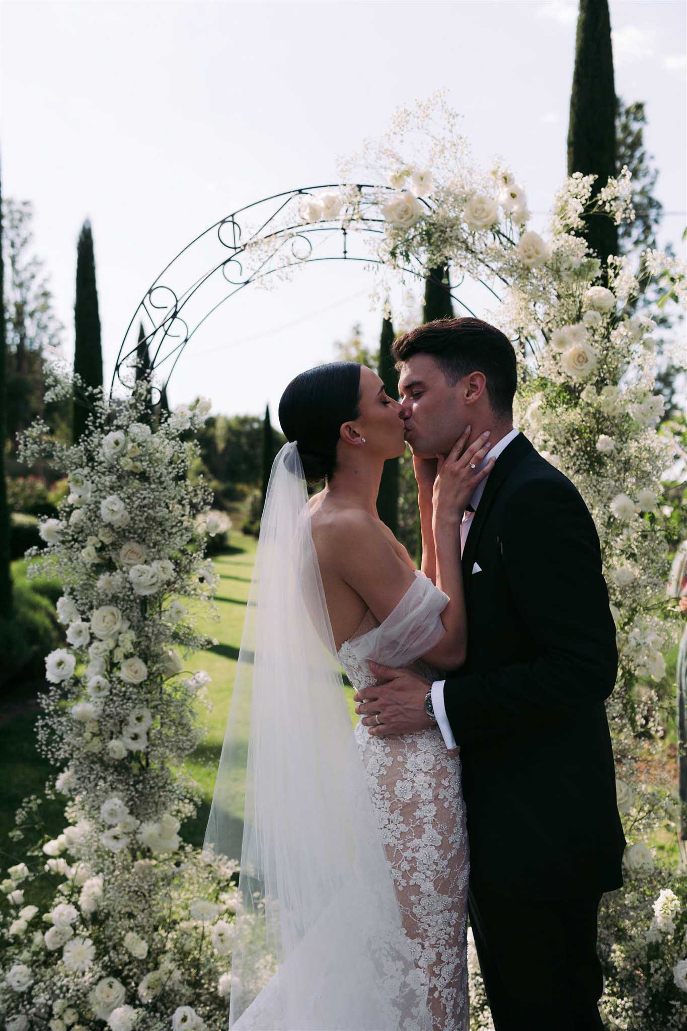 Bride and groom share first kiss under metal arch decorated with white roses and baby's breath in garden ceremony