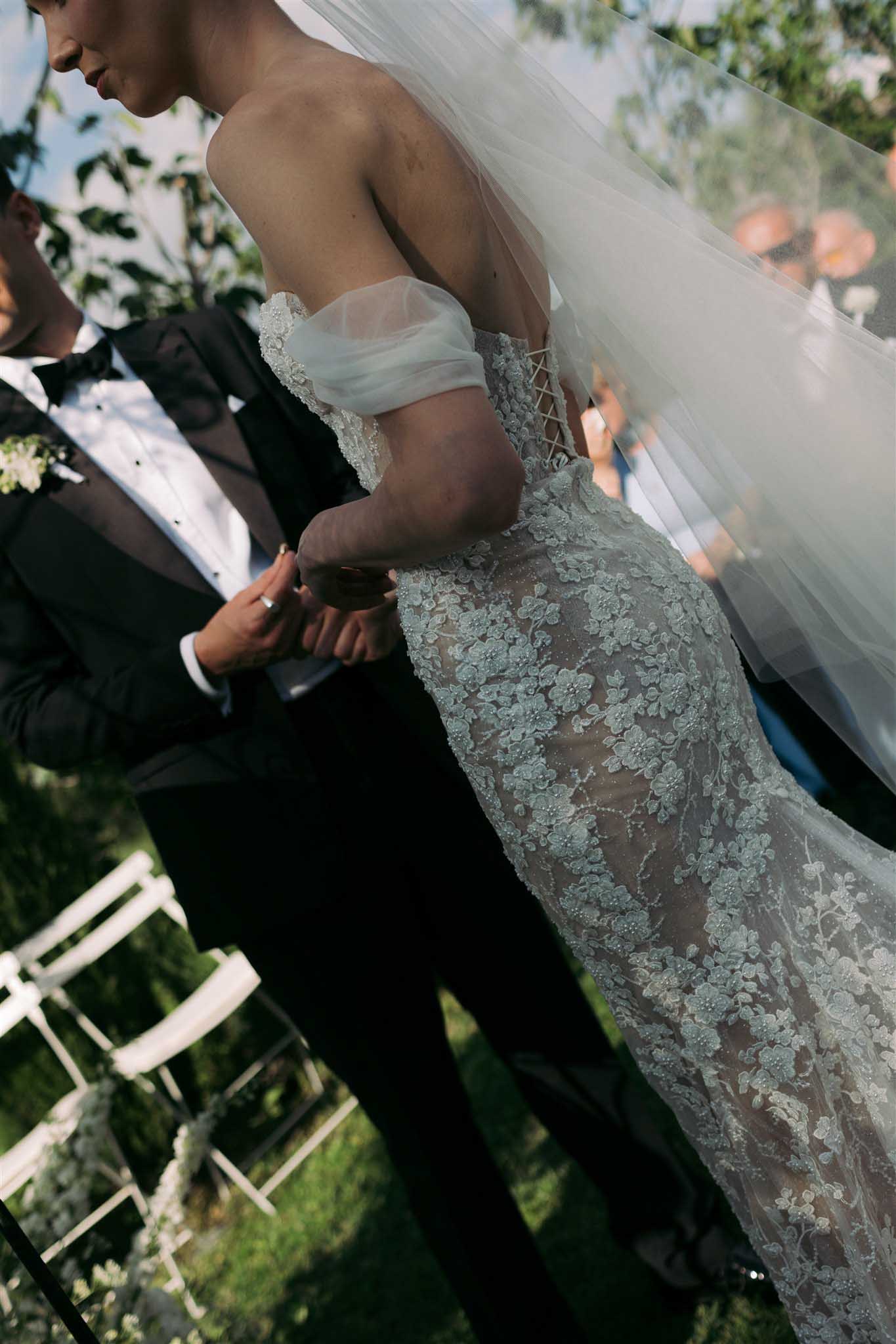 Bride in off-shoulder ivory lace gown with floral appliqués and voluminous veil during the outdoor ceremony, groom beside her.