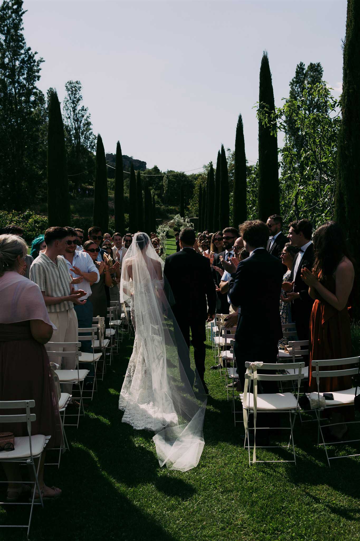 Bride in ivory lace gown walks aisle with groom as 80-100 standing guests applaud in cypress-lined garden ceremony