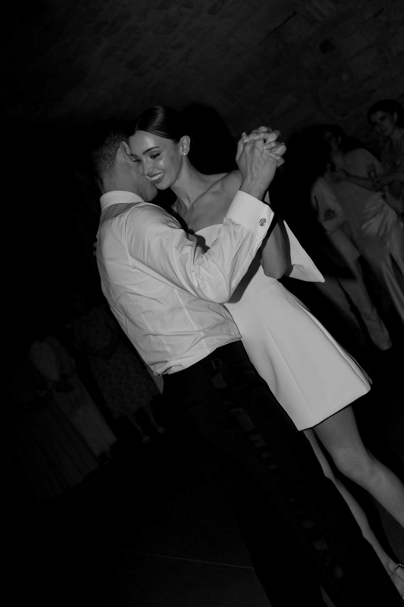 Black and white photo of groom lifting bride during first dance in off-shoulder white dress at indoor reception