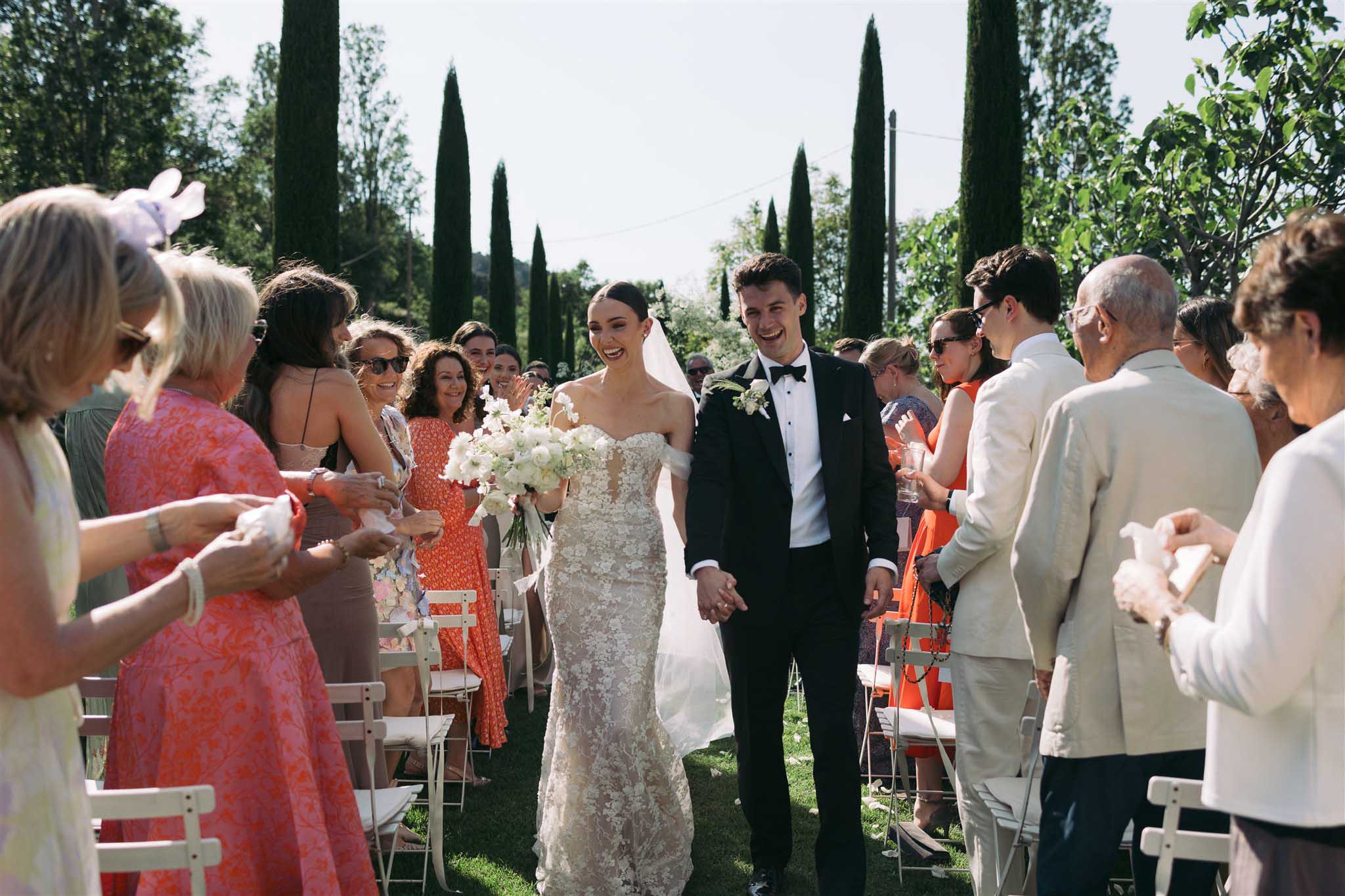 Bride and groom walk aisle after ceremony as guests toss white confetti, Mediterranean garden with cypress trees