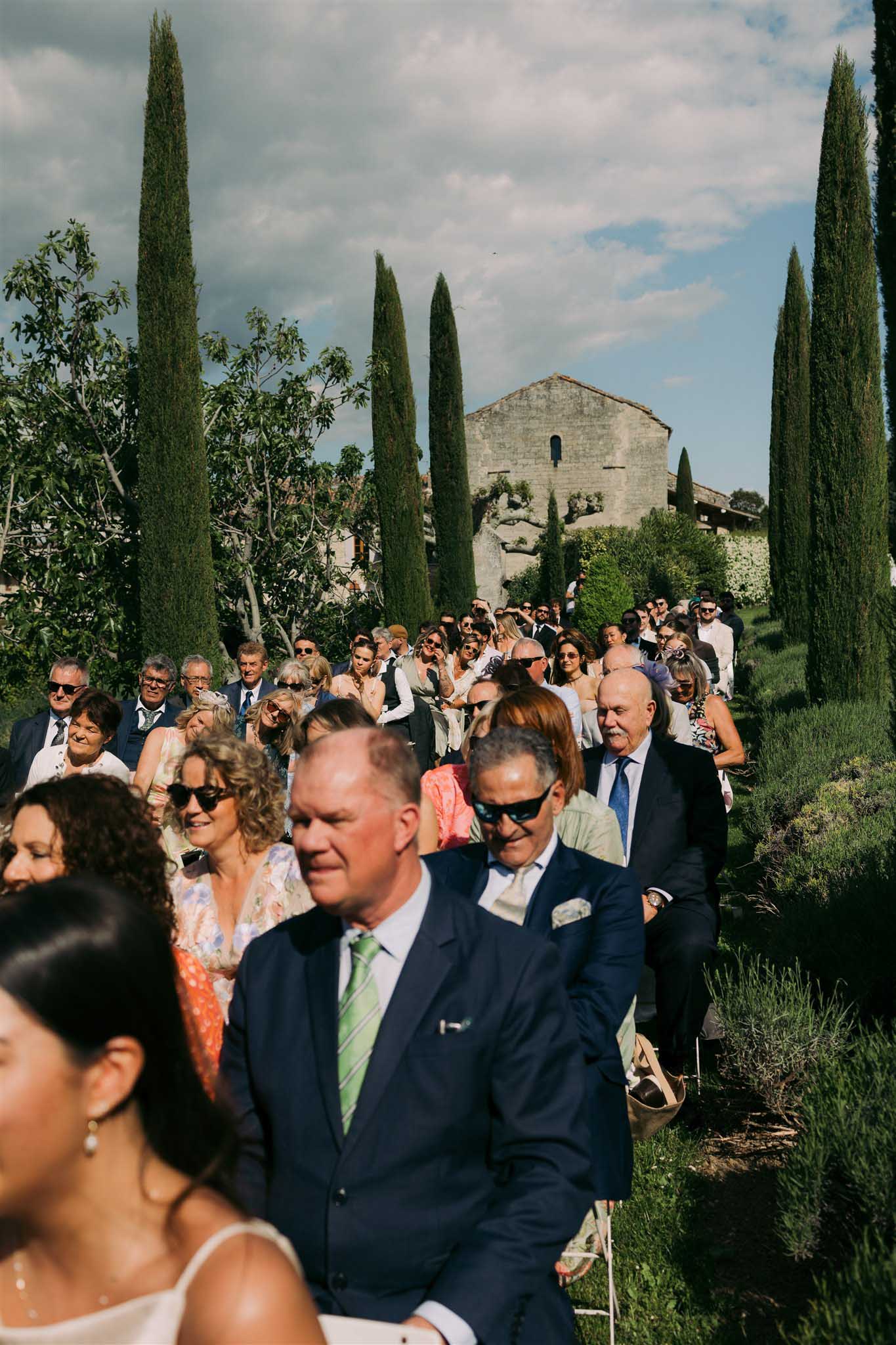 Wedding guests in formal attire walk along a tree-lined path toward a stone chapel for the ceremony