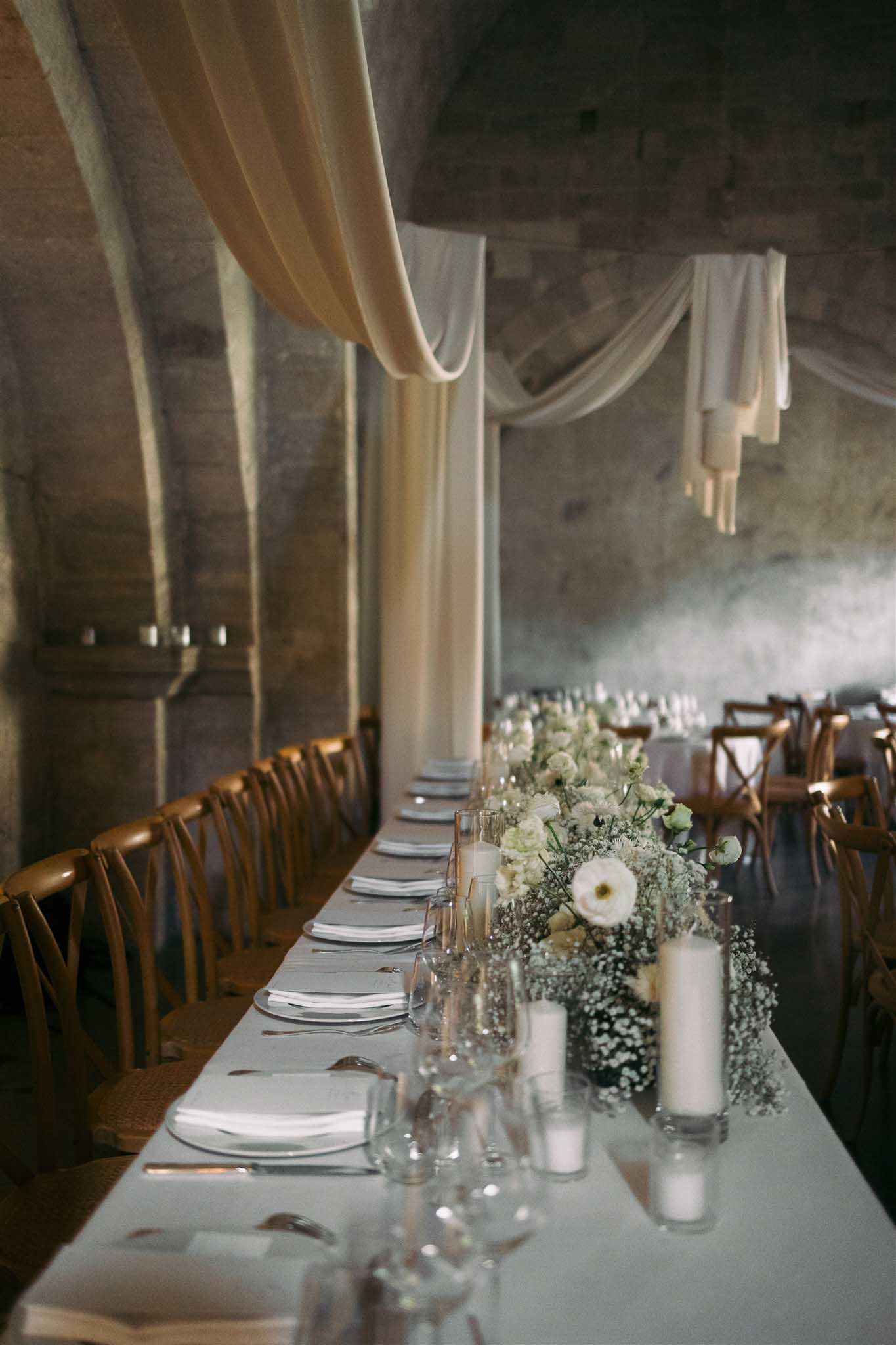 Indoor reception with long table draped in ivory fabric, white baby's breath runner, and cross-back chairs