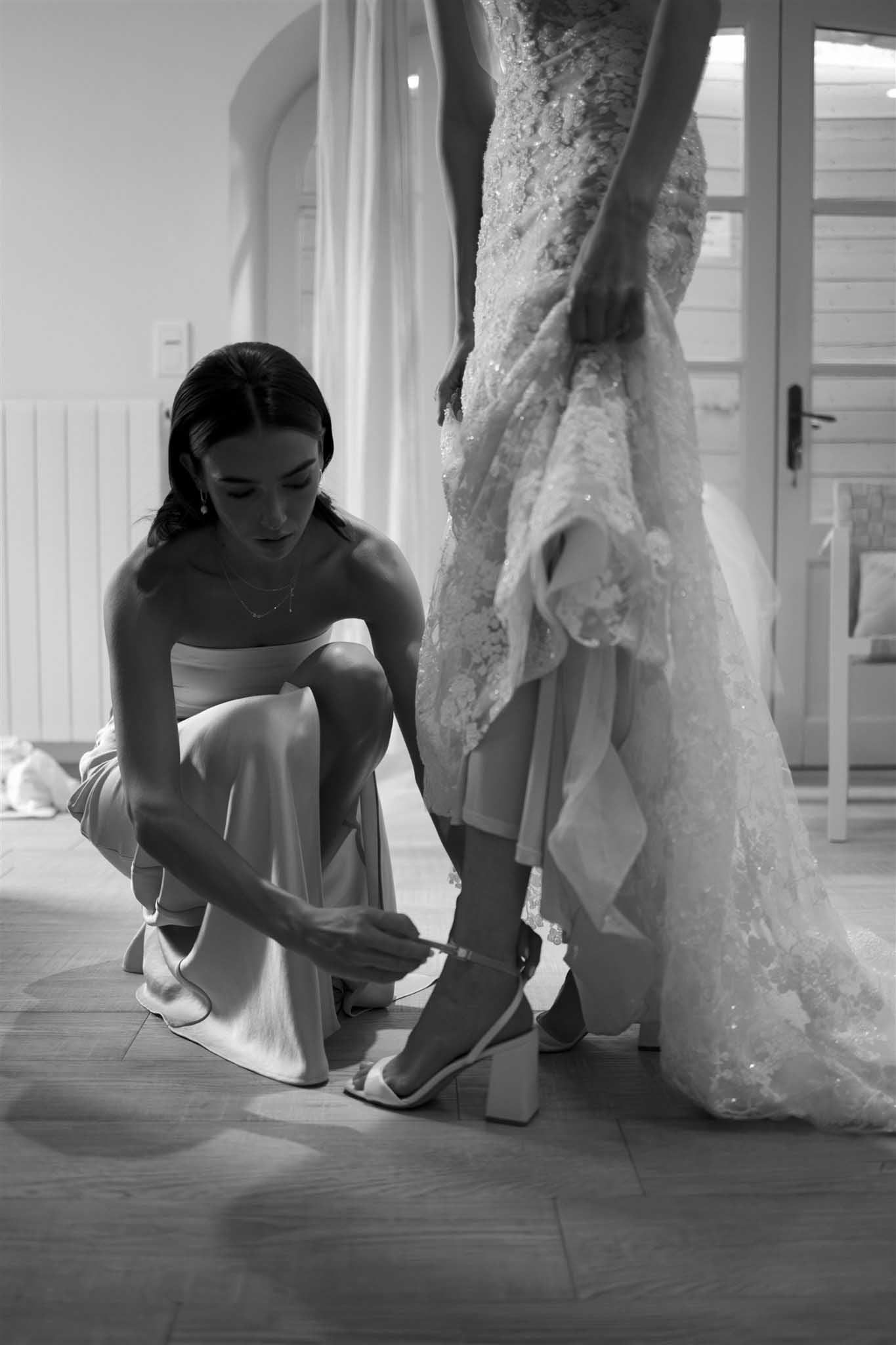Black and white photo of attendant helping bride adjust shoes showing beaded gown train detail