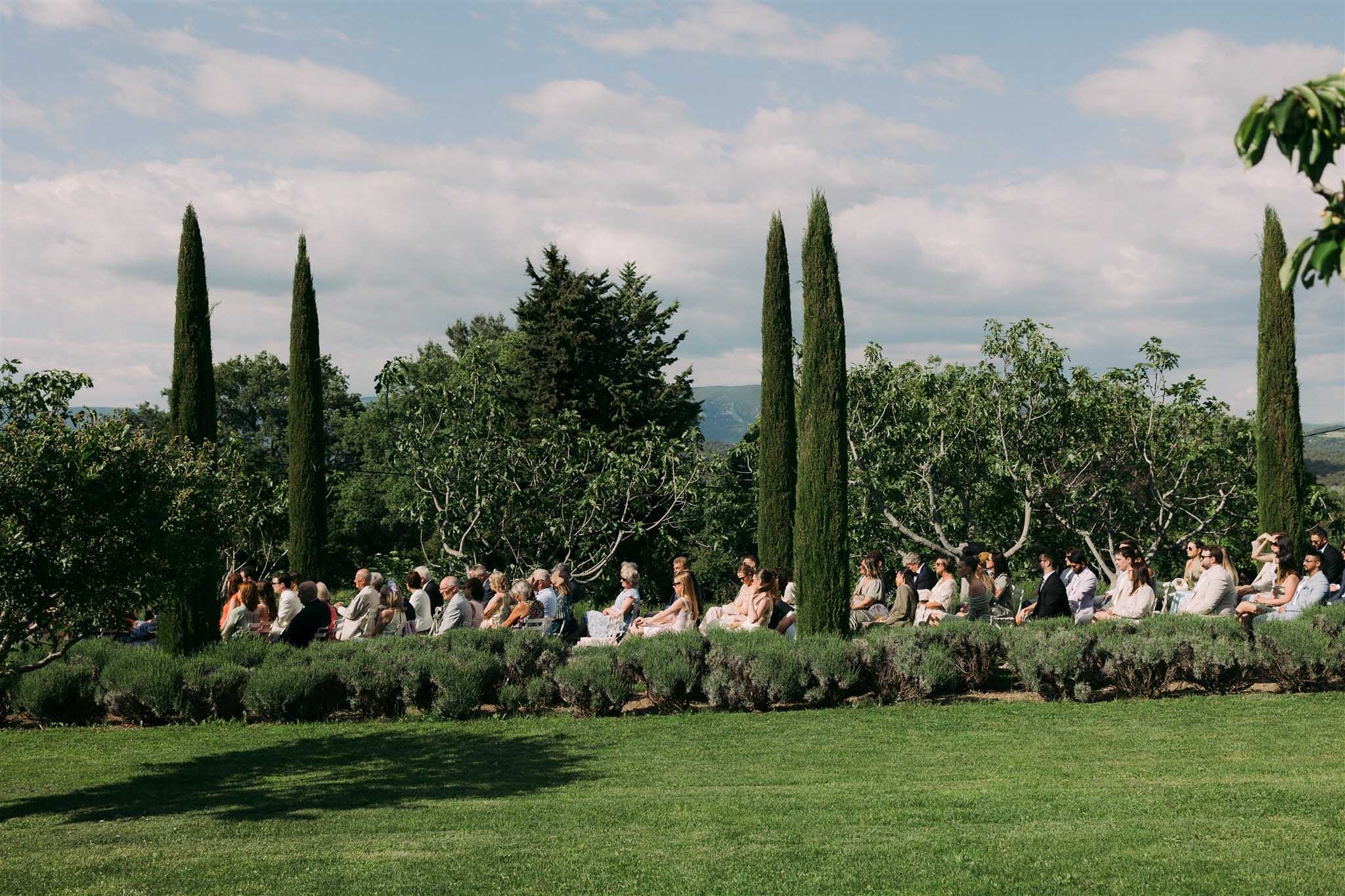 Guests seated at a long banquet table among olive and cypress trees in a Mediterranean garden reception setting