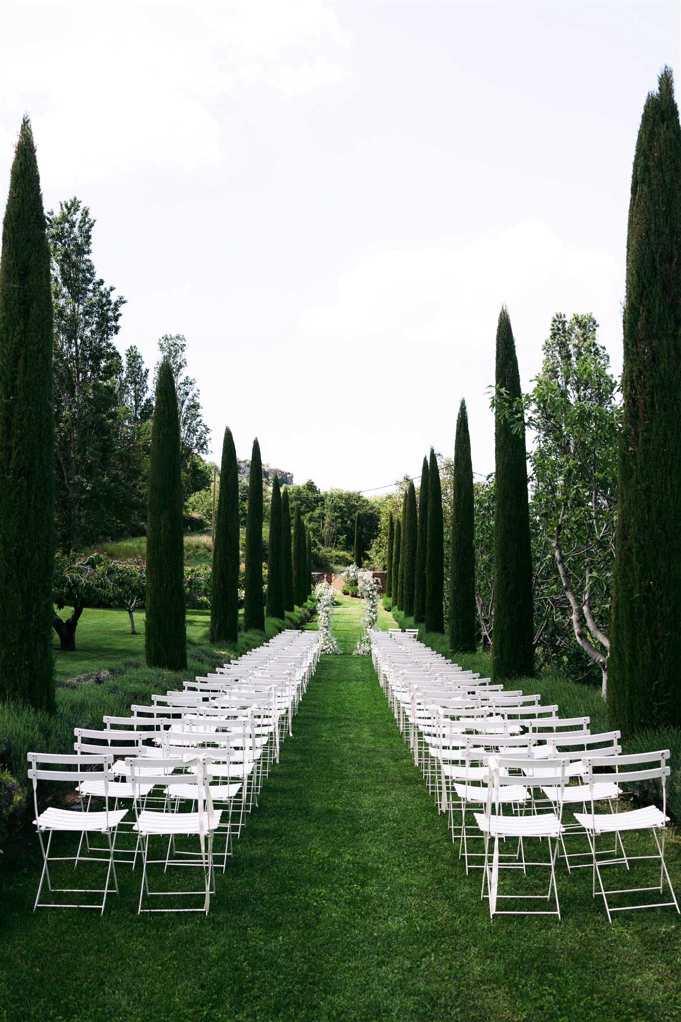 Wide-angle ceremony aisle with white folding chairs on manicured lawn, flanked by tall Italian cypress trees, formal garden