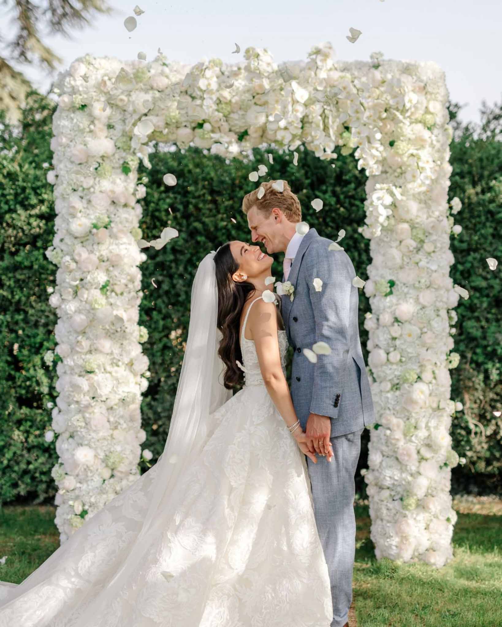 Bride and groom kissing under white hydrangea and rose arch with petals falling around them