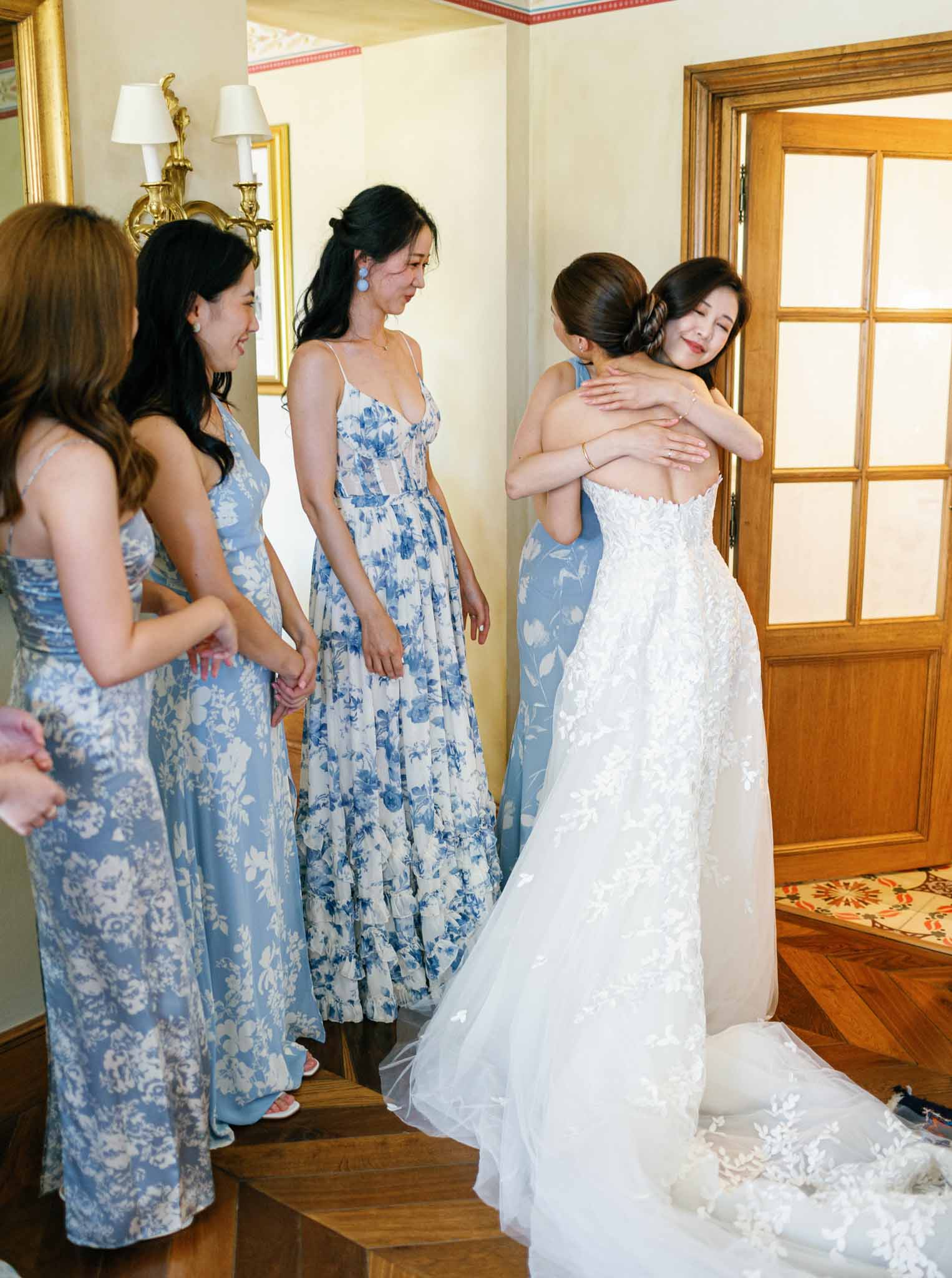 Bride in white beaded lace gown embracing bridesmaid while three attendants in blue floral dresses look on