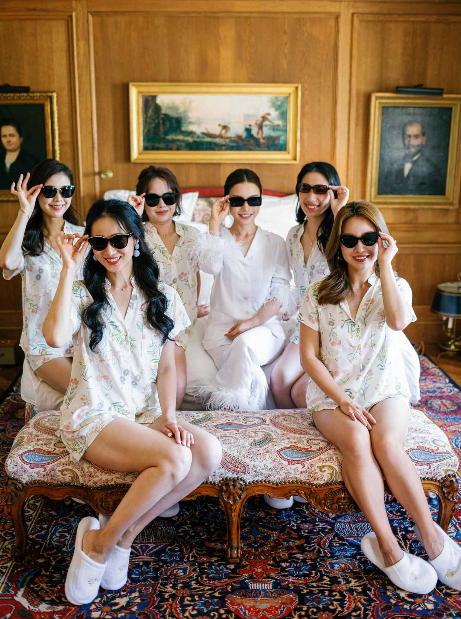 Bride and six bridesmaids in matching floral robes and black sunglasses posing playfully on ornate bed during preparations