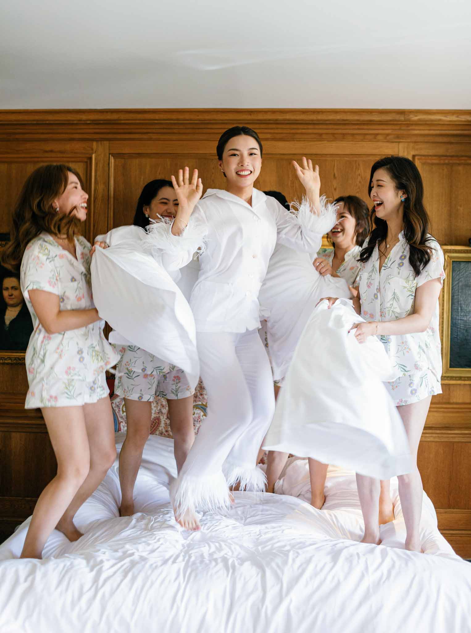 Bride jumping on bed with five bridesmaids in matching floral robes during pre-ceremony celebrations