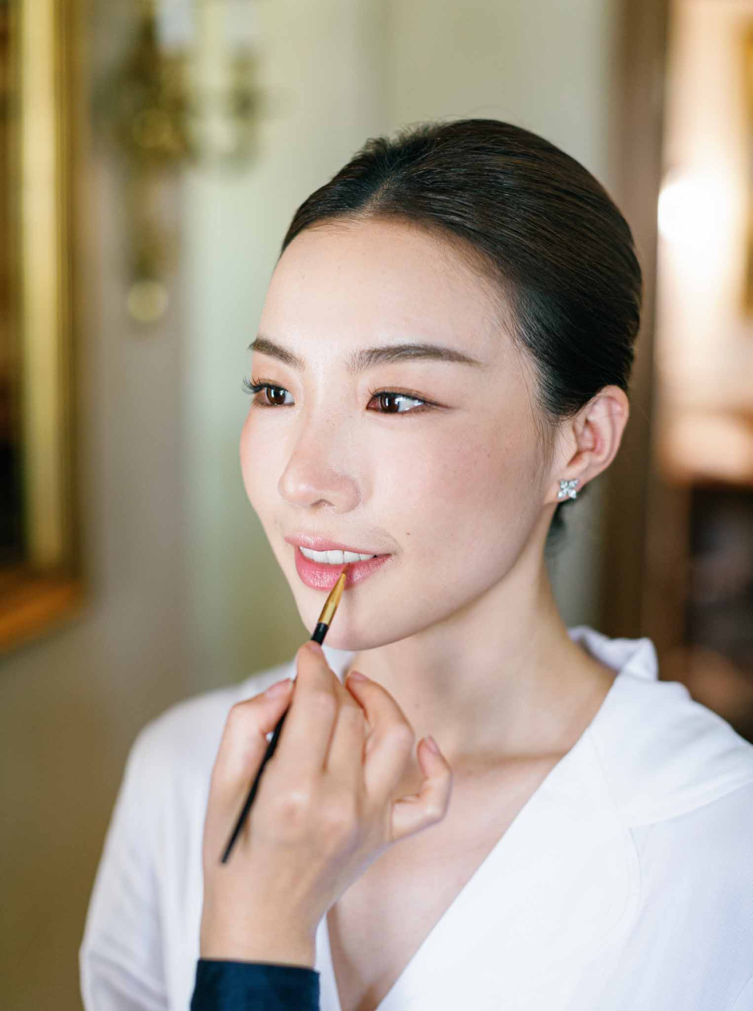 Bride having coral lipstick applied during makeup session in cream and gold wedding preparation room