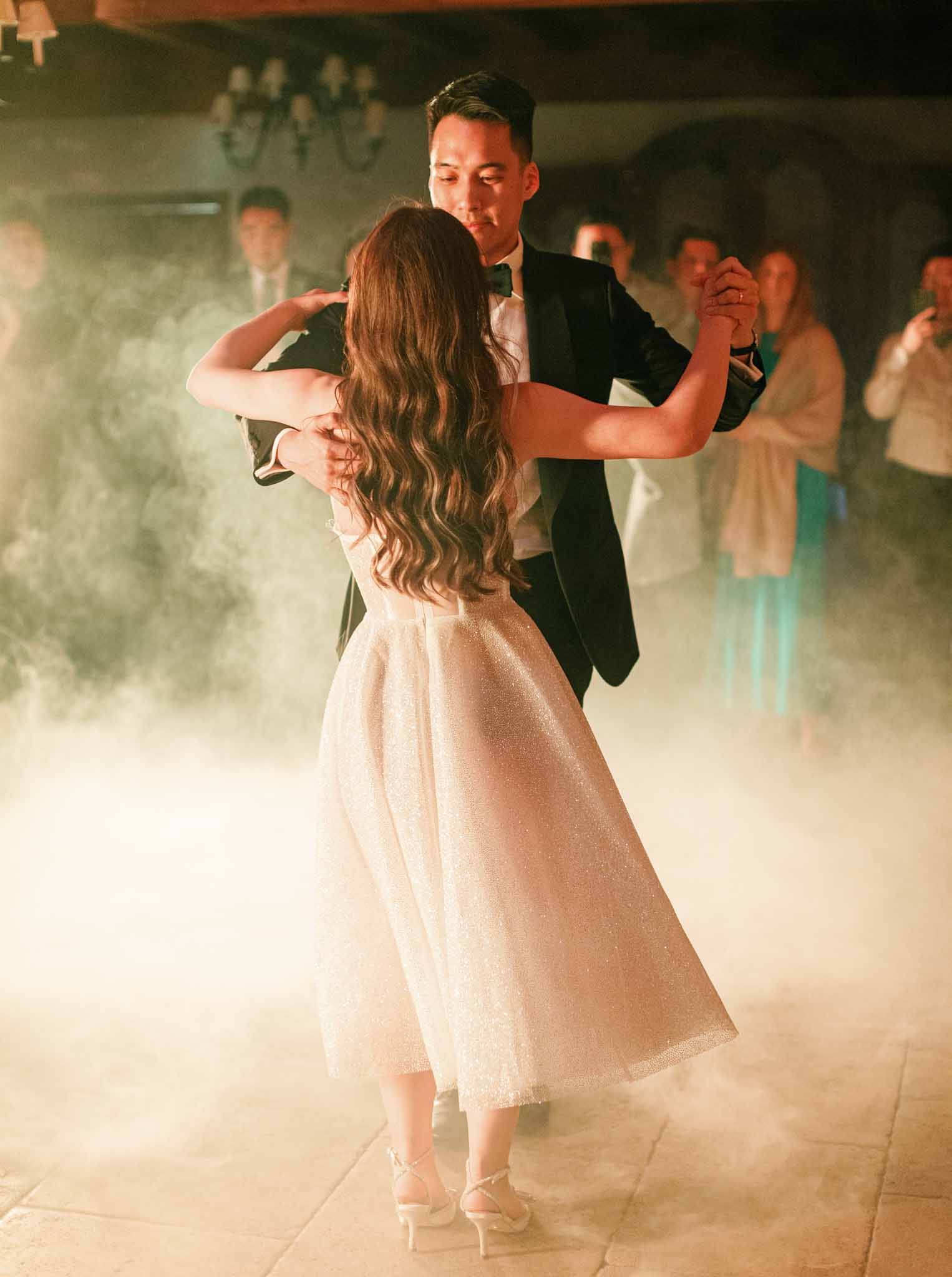 Bride in sequined champagne dress and groom in tuxedo sharing first dance surrounded by dry ice fog