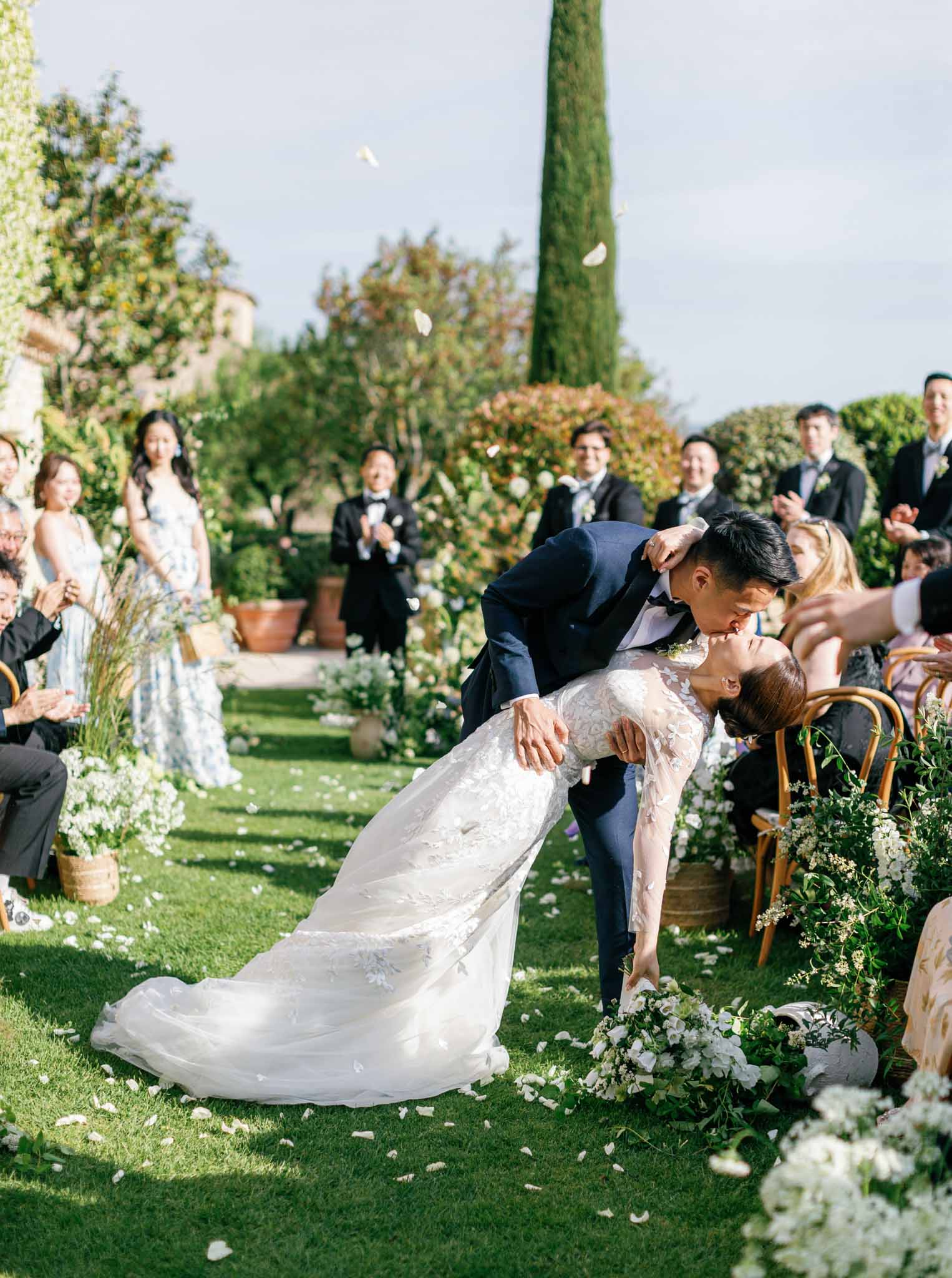 Groom dips bride for their first kiss at the end of an outdoor garden ceremony lined with white flowers