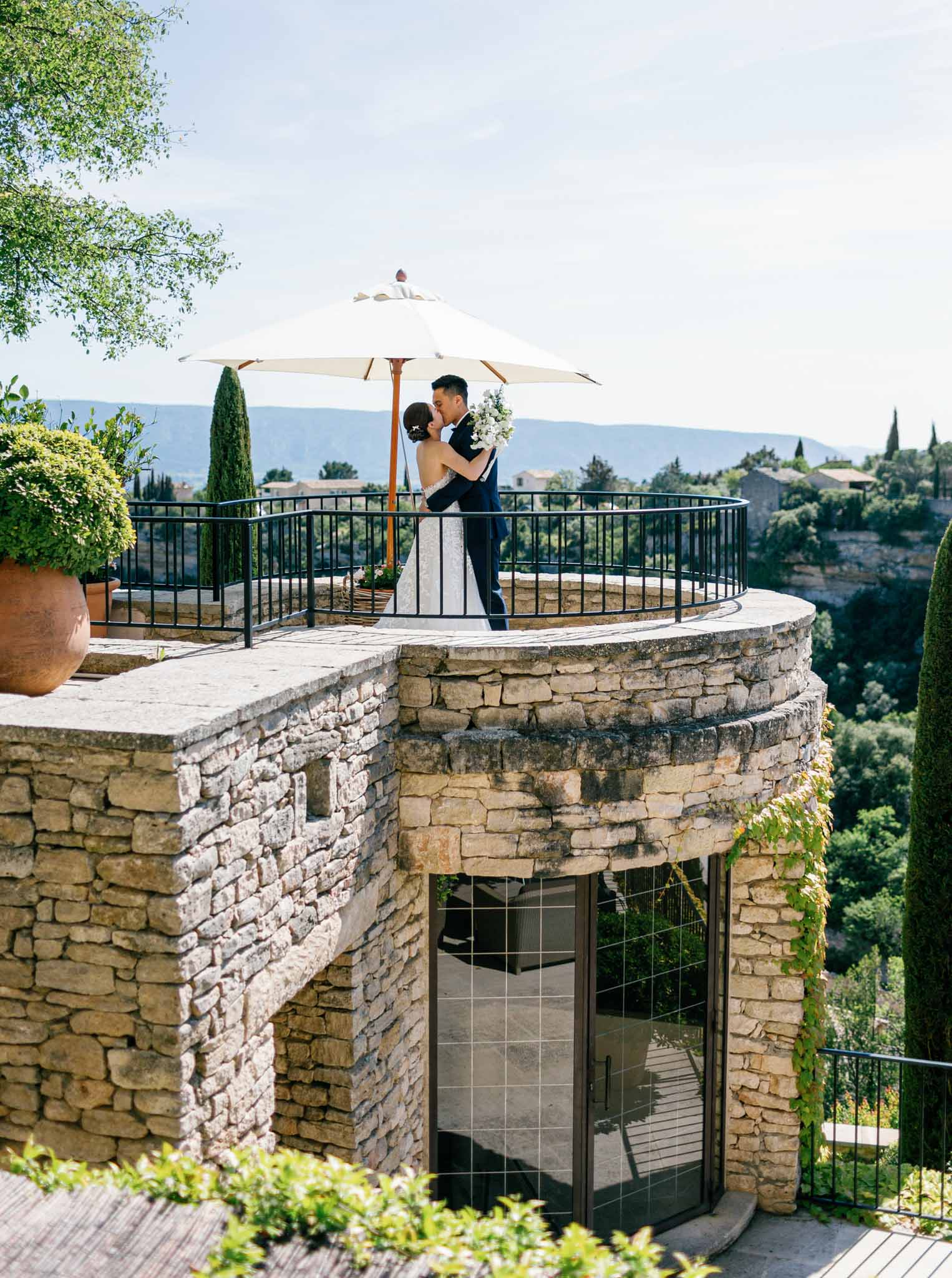 Couple on stone circular terrace with umbrella overlooking hillside landscape at Mediterranean-style venue with cypress trees