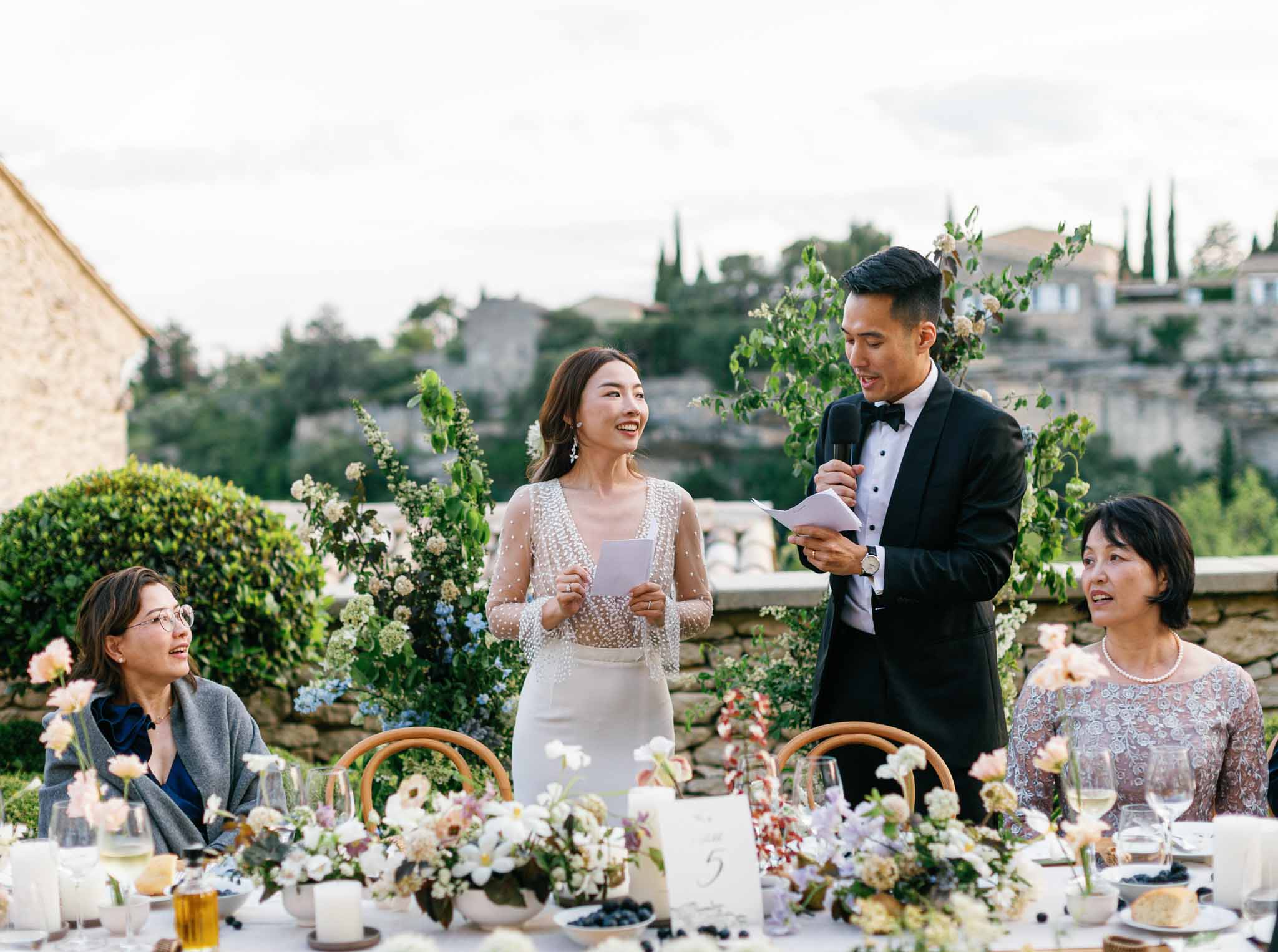 Groom in tuxedo delivering speech with microphone at outdoor reception table, bride seated smiling with Tuscan hillside backdrop