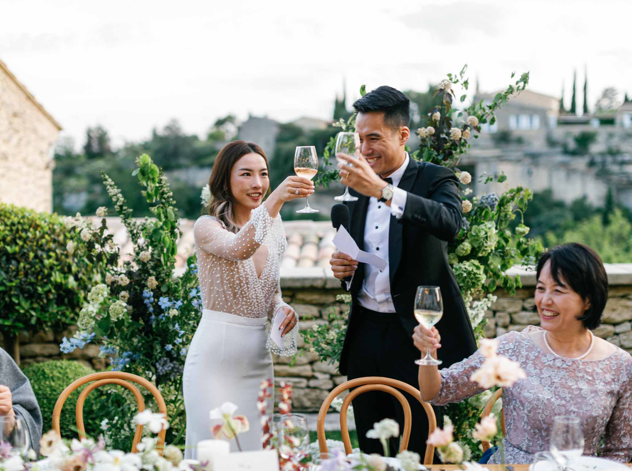 Couple toasting on outdoor terrace with hillside village behind, tall white and pale blue florals, guests nearby