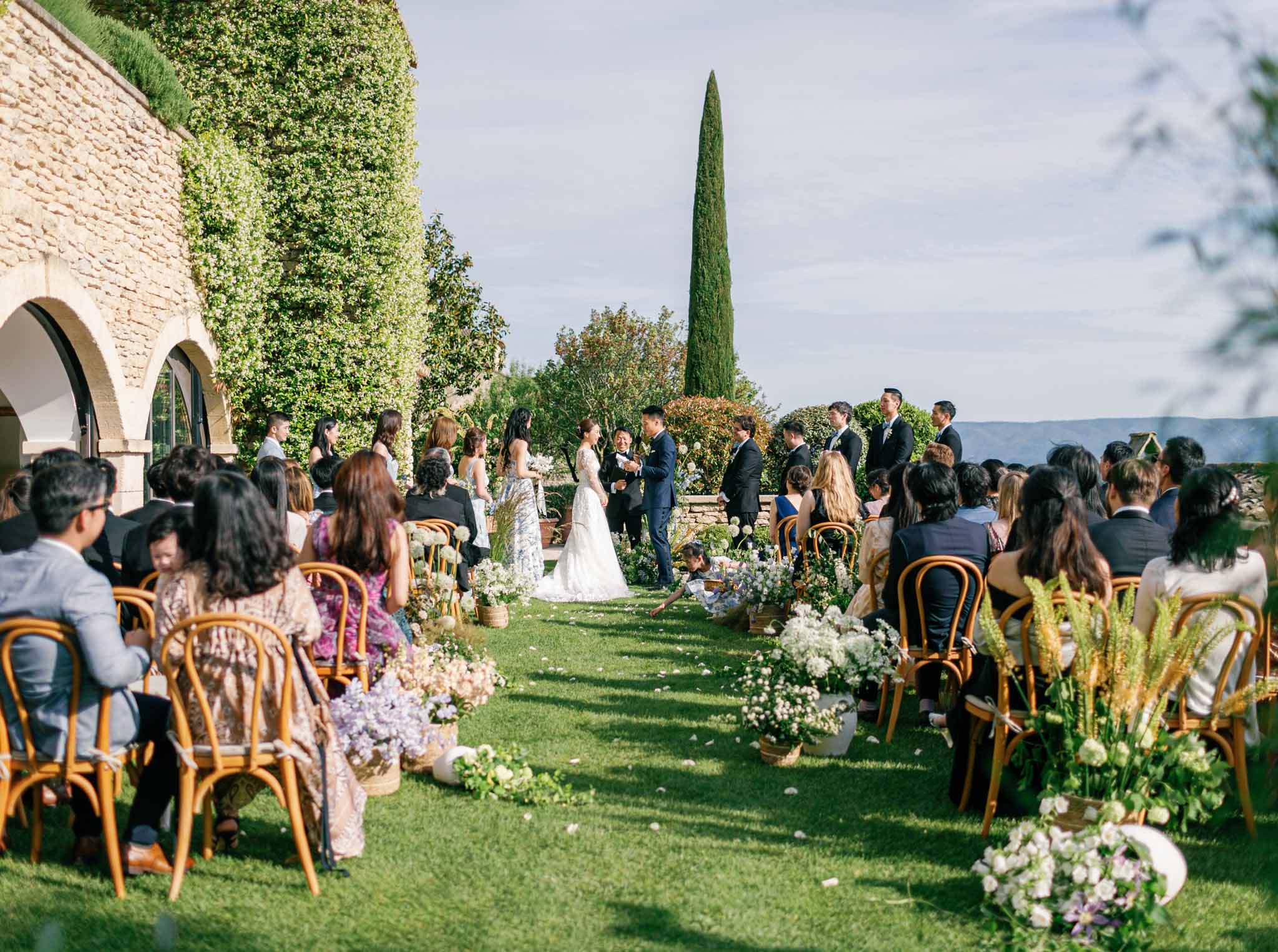 Outdoor wedding ceremony in manicured garden with guests seated on wooden chairs, ivy-covered stone building and cypress tree backdrop