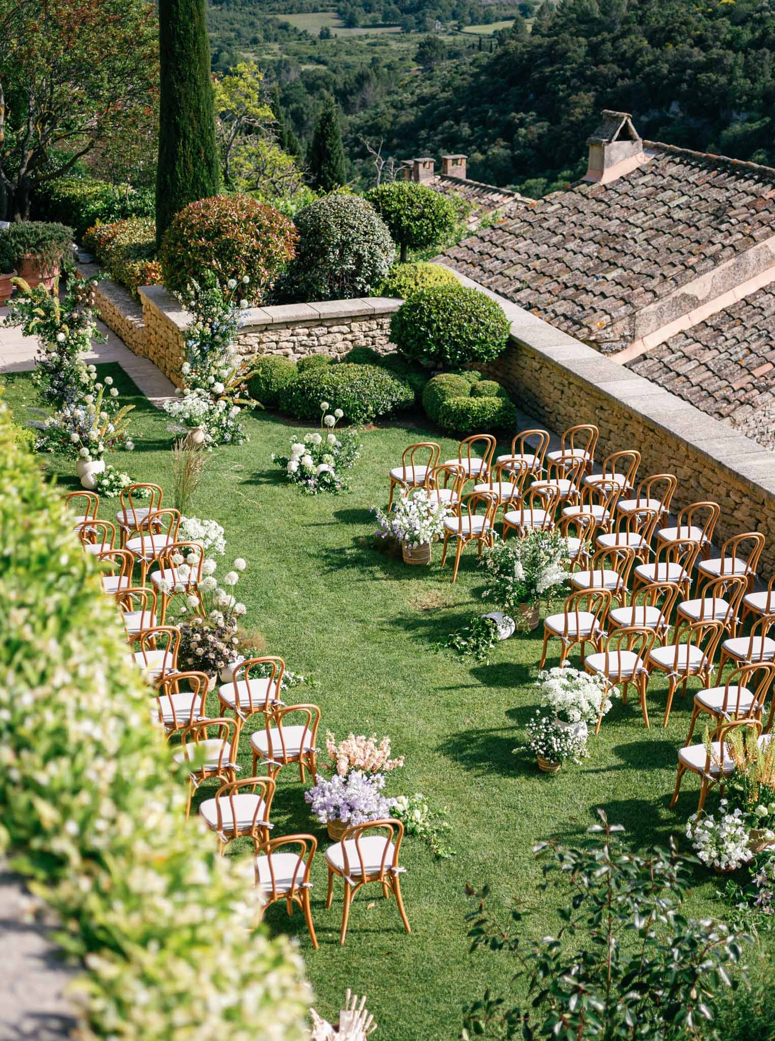 Outdoor ceremony setup on manicured lawn with bentwood chairs and white floral planters at stone-walled Provencal venue