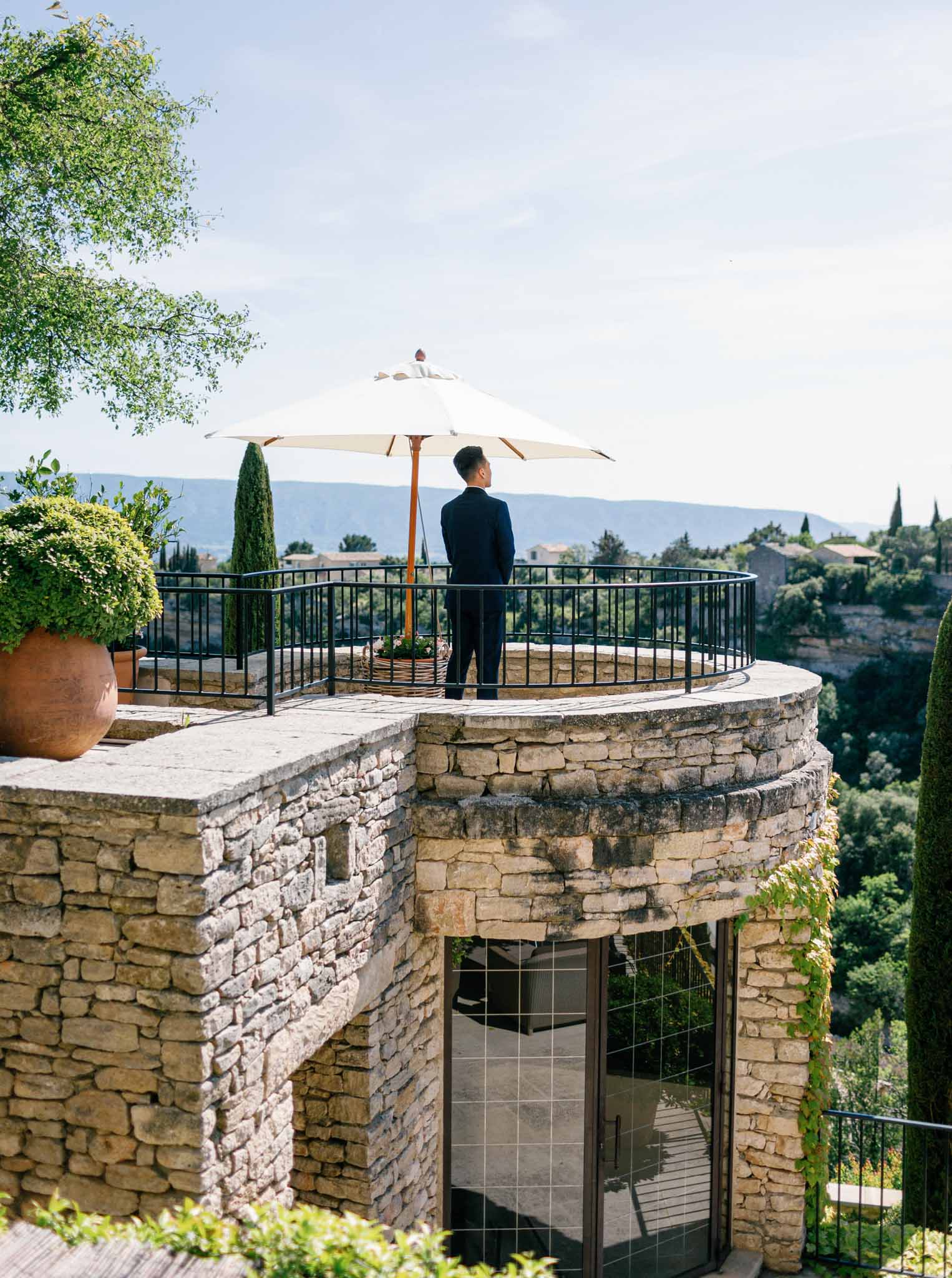 Groom standing on circular stone terrace overlooking rolling hills and cypress trees at Mediterranean venue
