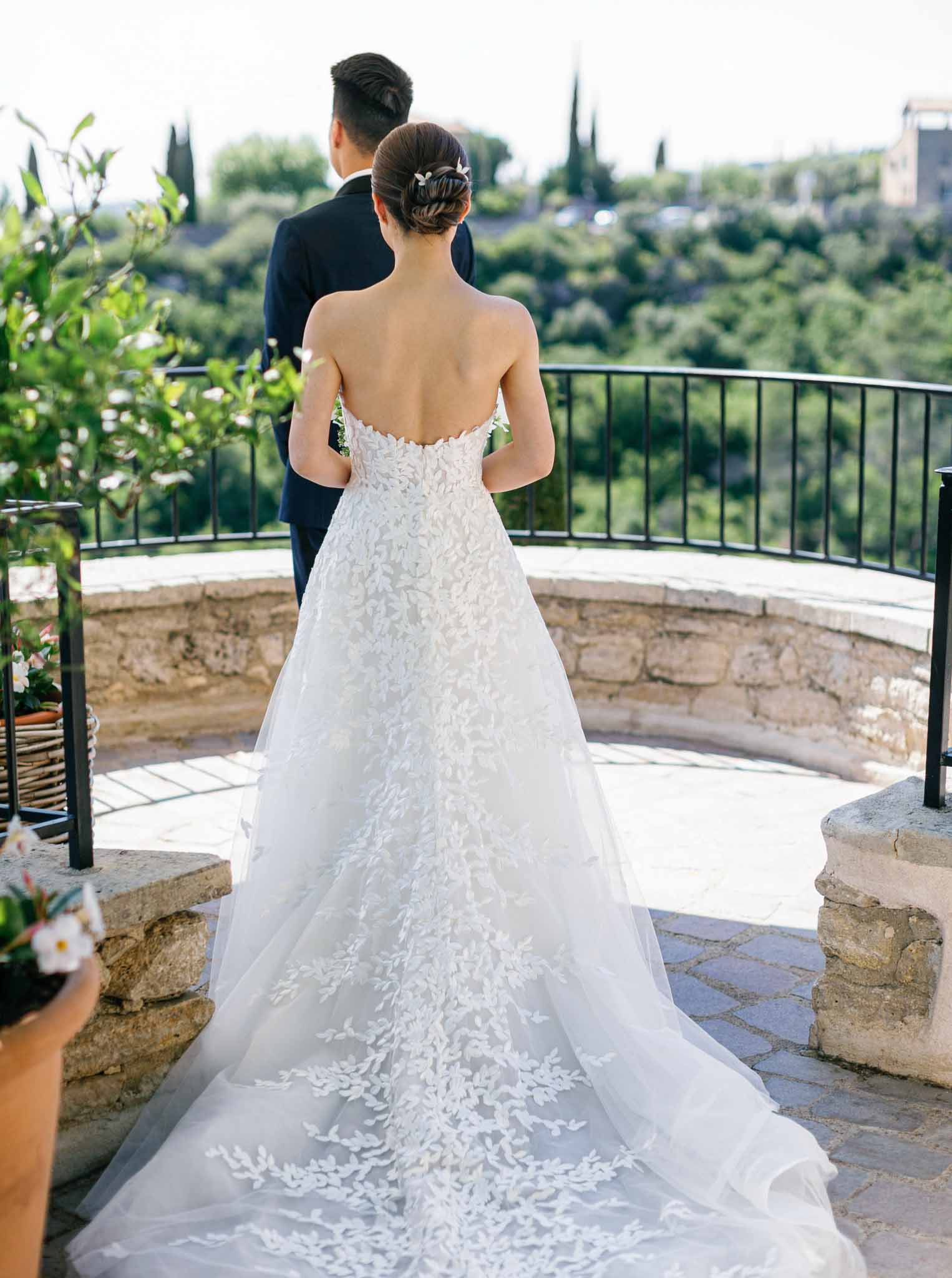 Rear-view portrait of bride in lace appliqué A-line gown and groom in navy suit on stone terrace overlooking Mediterranean hills