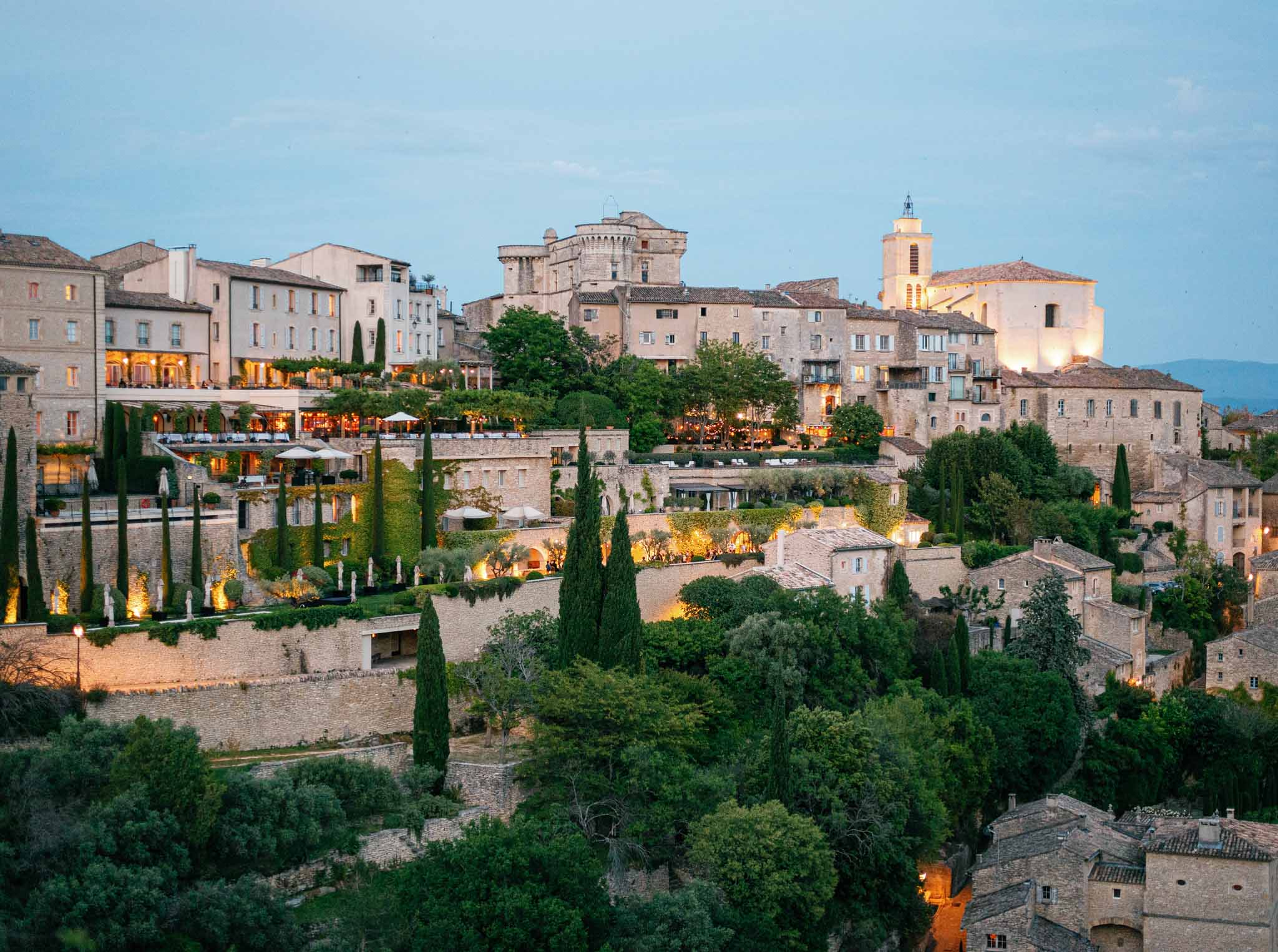 Provençal hilltop stone village at twilight with warm golden lighting, church bell tower, and cypress-lined terraces