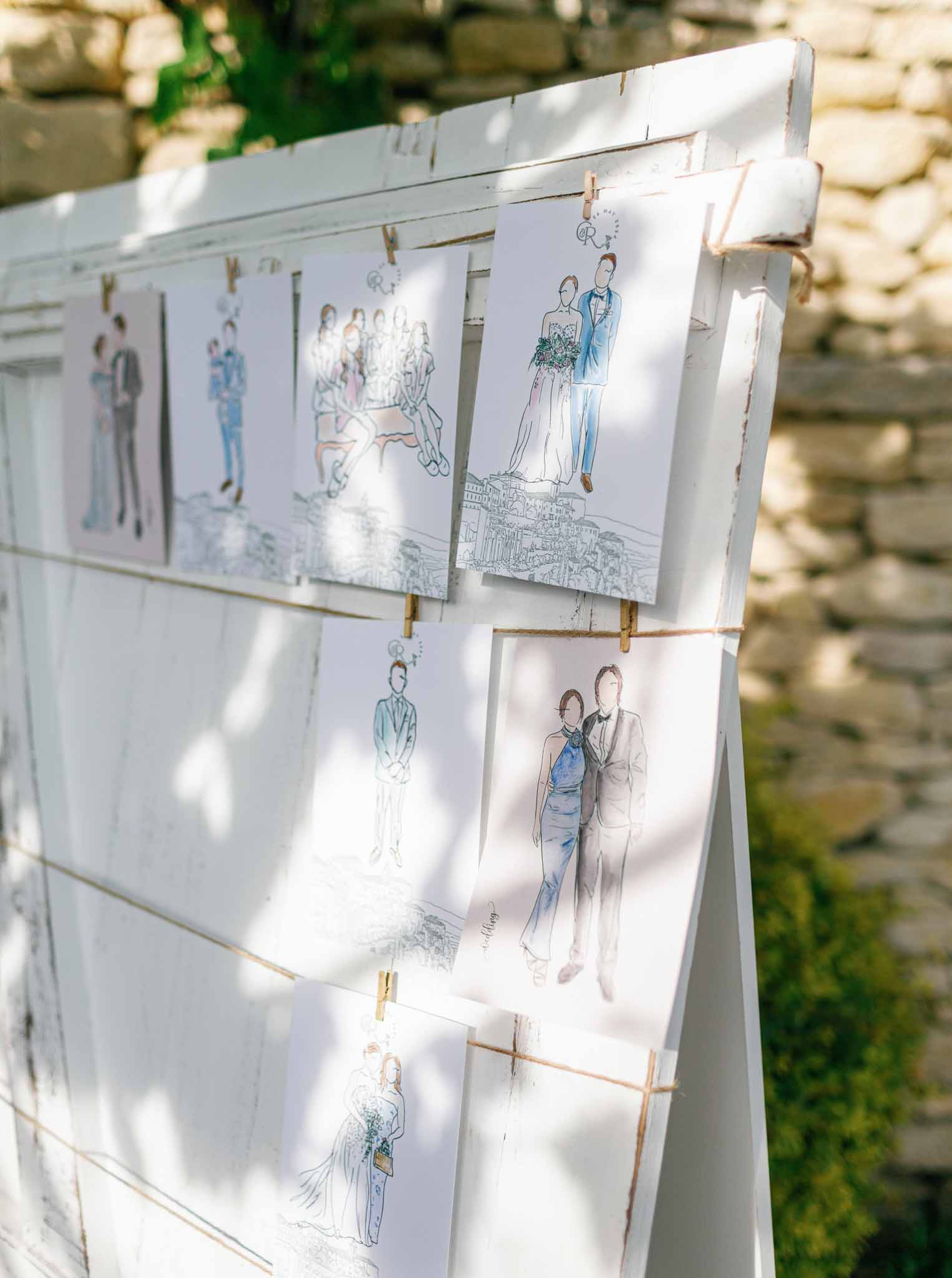 Watercolor fashion sketches of wedding party clipped to wooden display board against ivy-covered stone wall