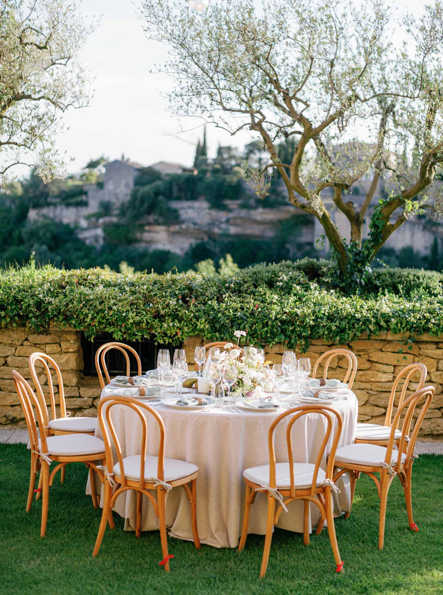 Round reception table with cream linen, bentwood chairs, white and blush centerpiece on a lawn in front of a Mediterranean stone wall.