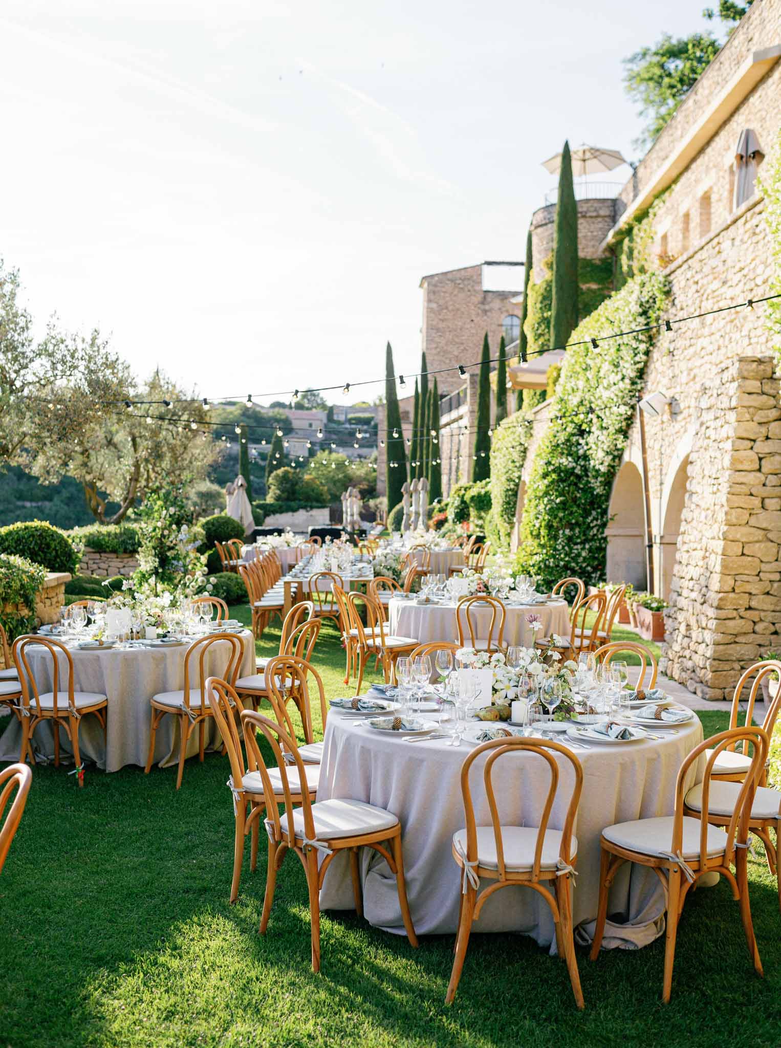Outdoor reception with round tables, grey linens, and white rose centerpieces beside ivy-covered stone villa