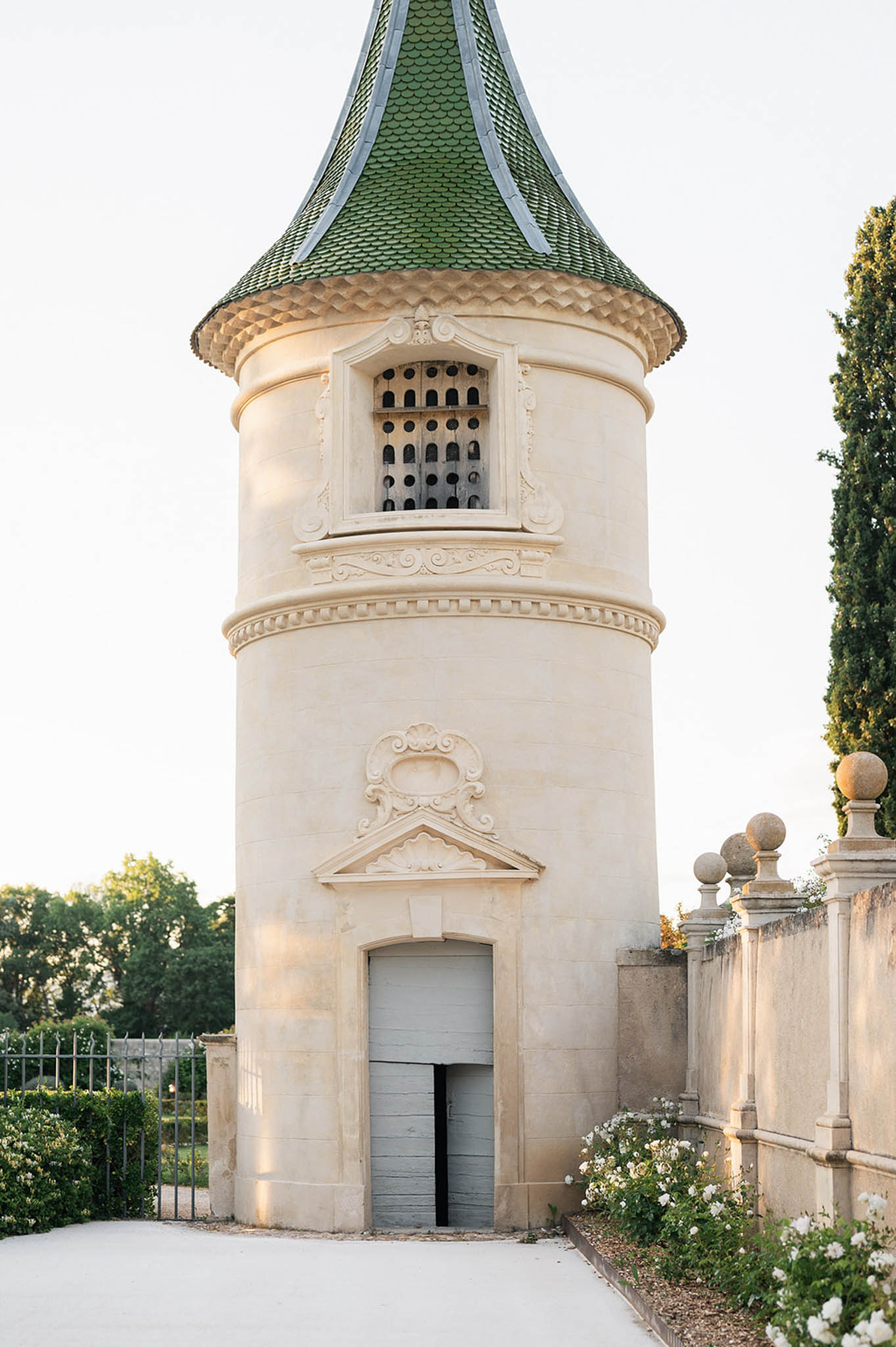 Circular stone gatehouse tower with baroque carvings, green glazed tile roof, and white roses at base