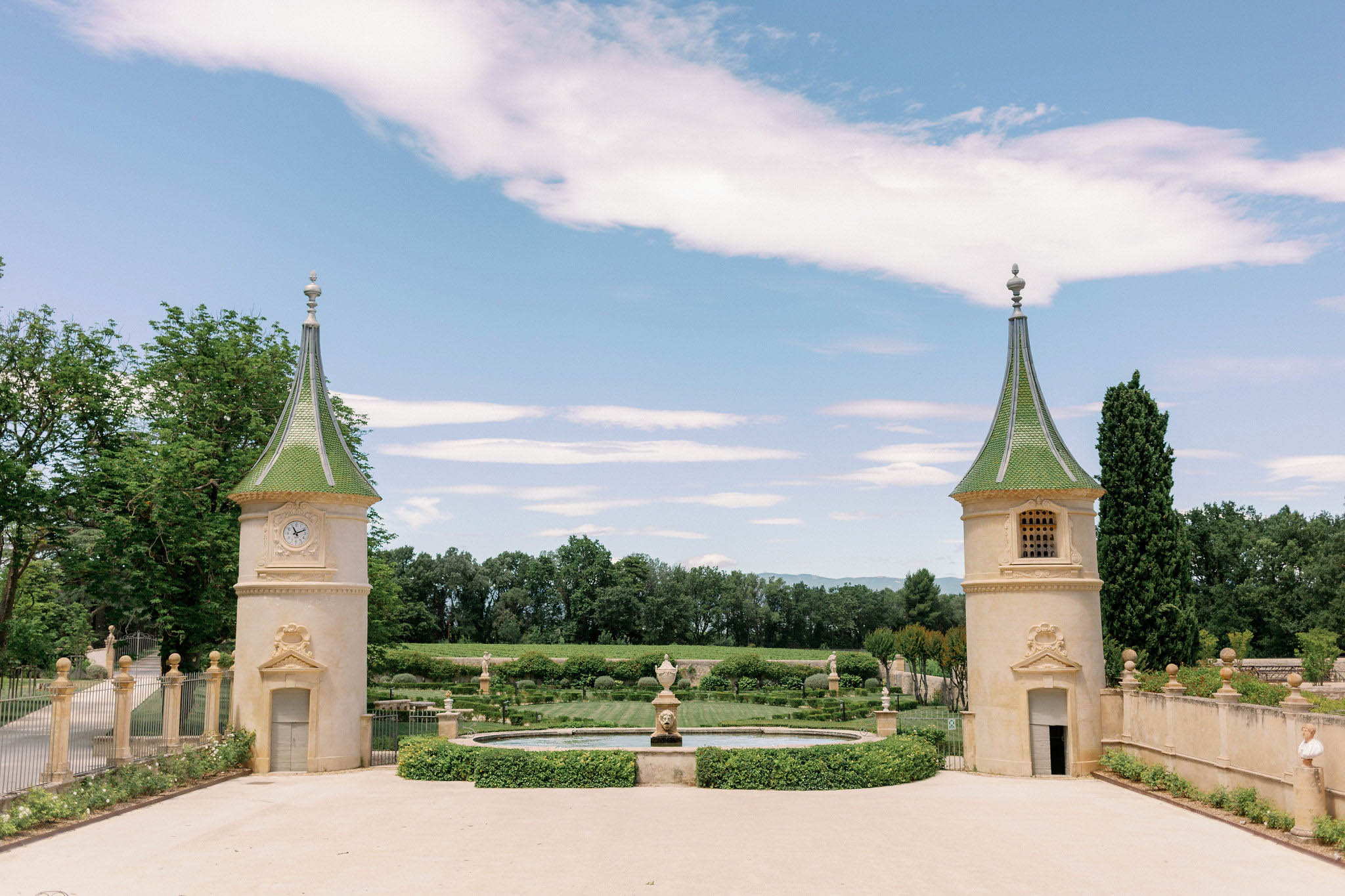 French chateau entrance with twin stone pavilion towers, formal boxwood garden, circular fountain, and vineyard beyond