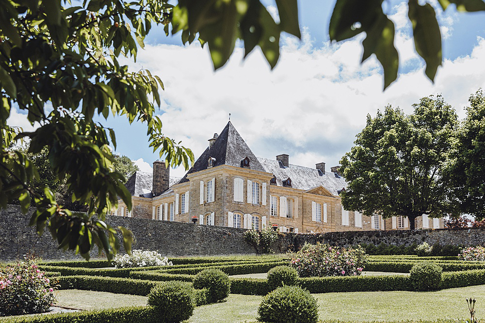 A wide exterior shot of a French château viewed from its formal garden, with no people present. The building features classic French architecture with golden limestone façades, steep slate roofs with pointed turrets, white shuttered windows, and multiple chimneys. In the foreground, a formal parterre garden is visible with neatly trimmed boxwood hedges arranged in geometric patterns, rounded topiary balls, and flowering rose bushes in pink and white. The image is framed by overhanging tree branches in the upper left corner. The composition is a medium-wide landscape shot taken from ground level within the garden. Potential venue feature image.