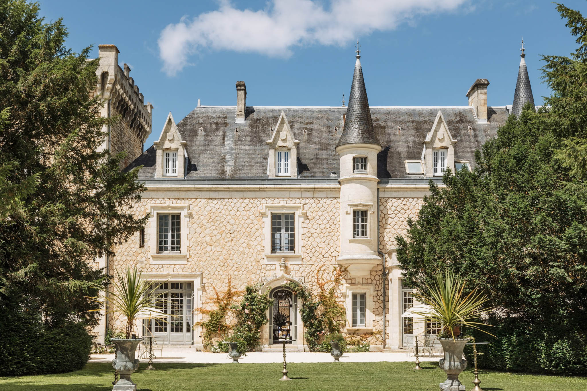 French chateau exterior with golden limestone facade, slate mansard roof, round turret, iron gate, and stone urns