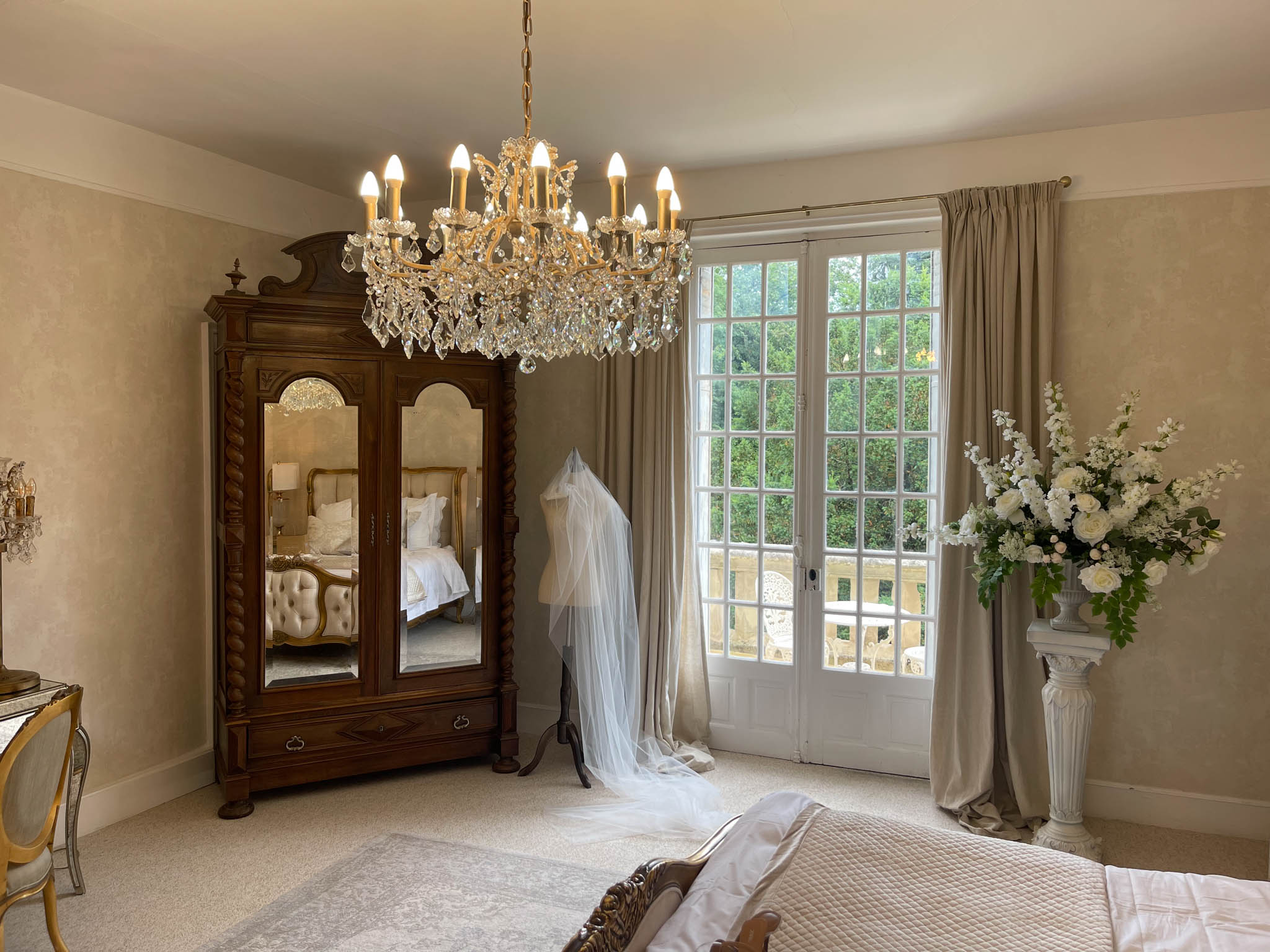 Chateau bridal suite with tulle veil on dress form white rose arrangement antique armoire and crystal chandelier
