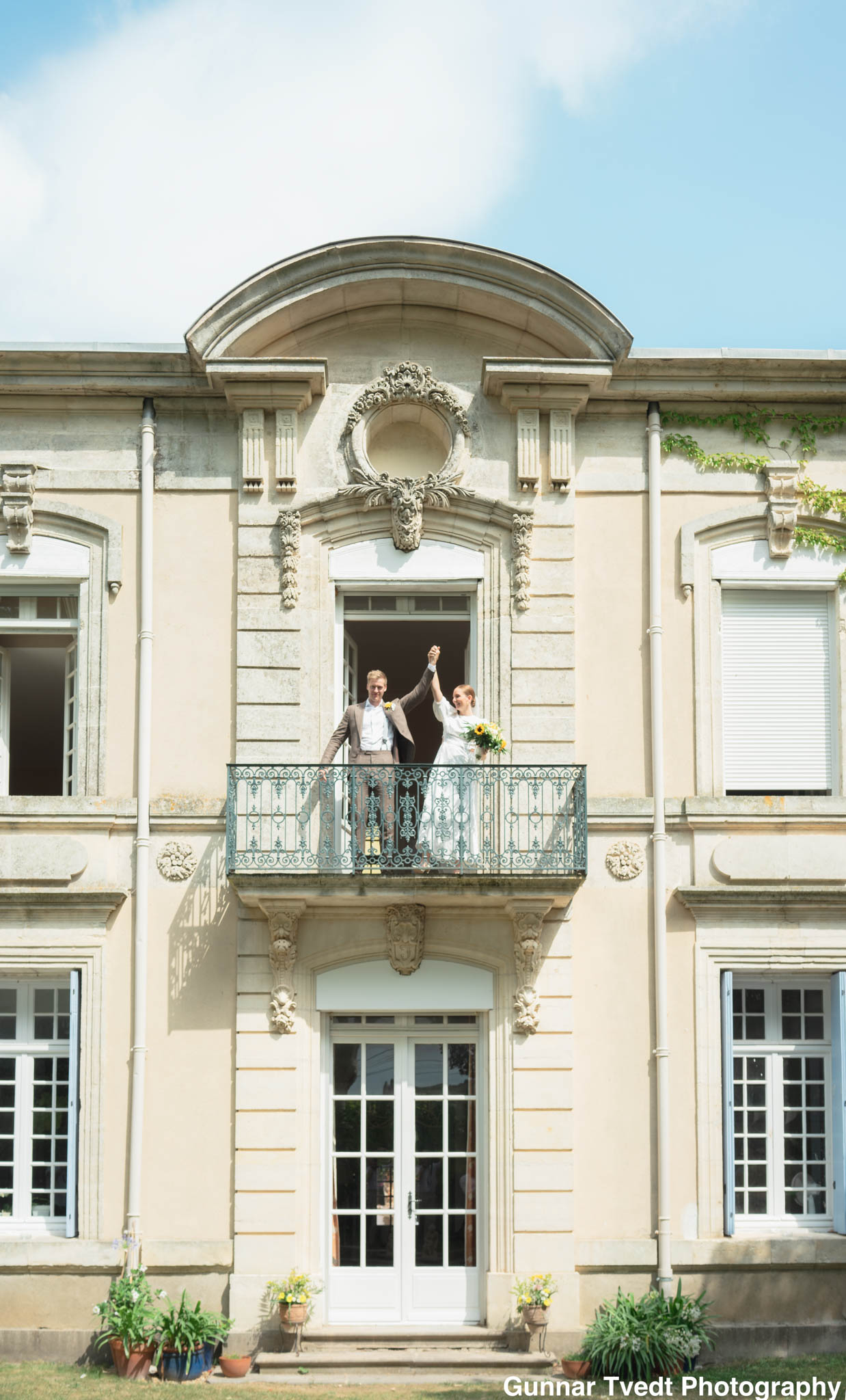 Bride and groom raise joined hands on ornate chateau balcony, bride holding sunflower bouquet