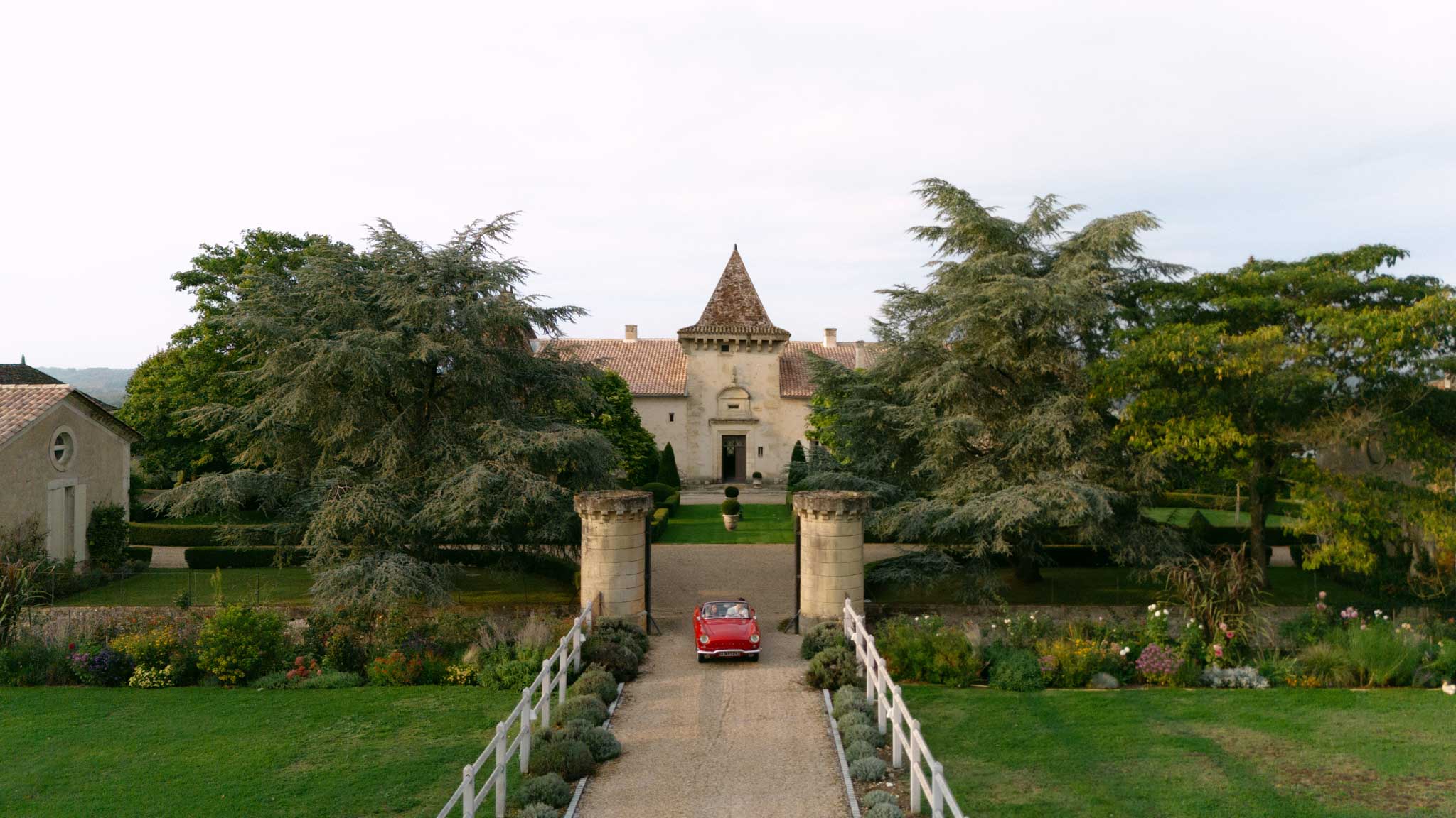 Vintage red convertible driving up gravel driveway toward French chateau with stone turret and flower-lined path