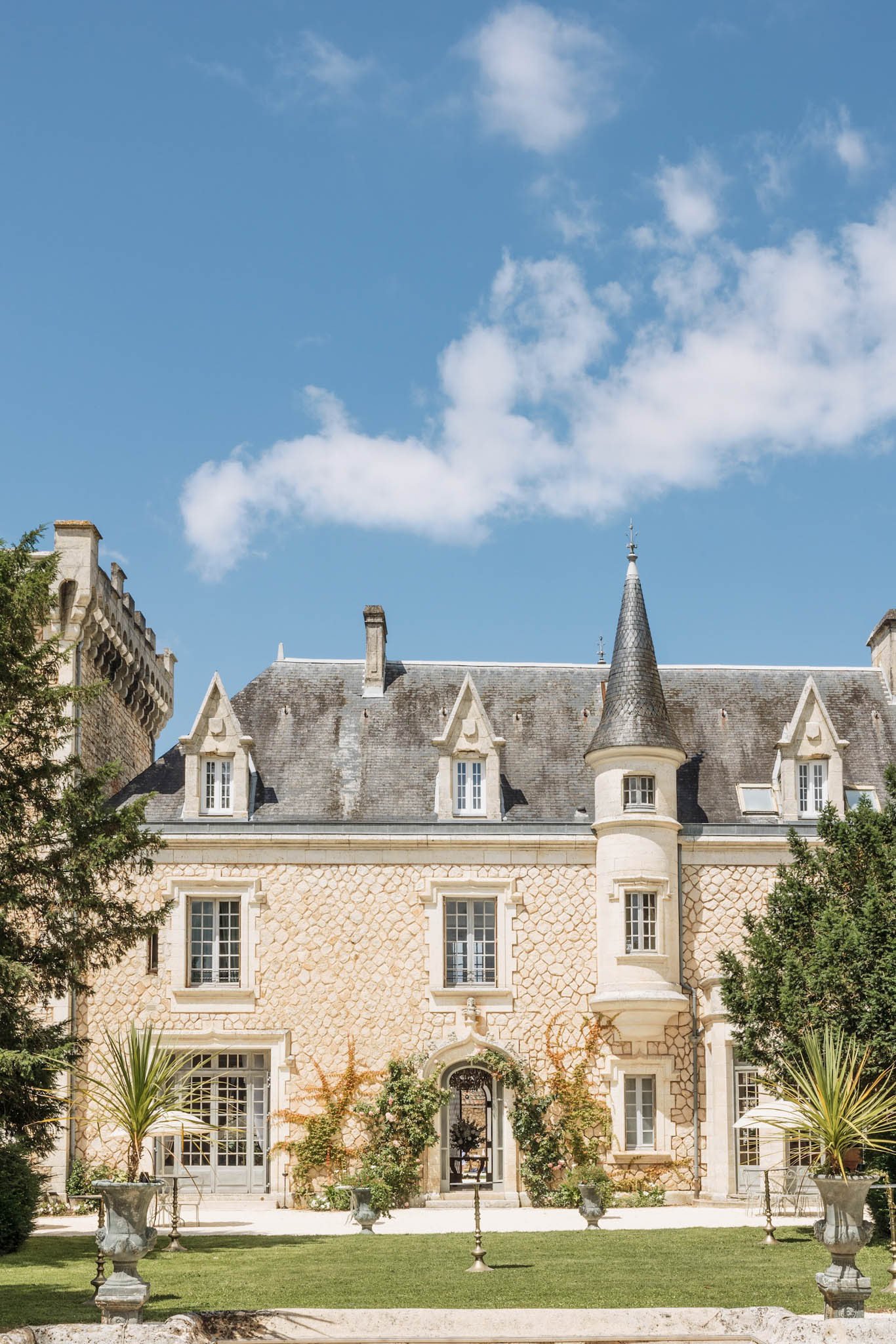 Golden limestone chateau with diamond-cut facade, mansard roof, and conical turret flanked by stone urns