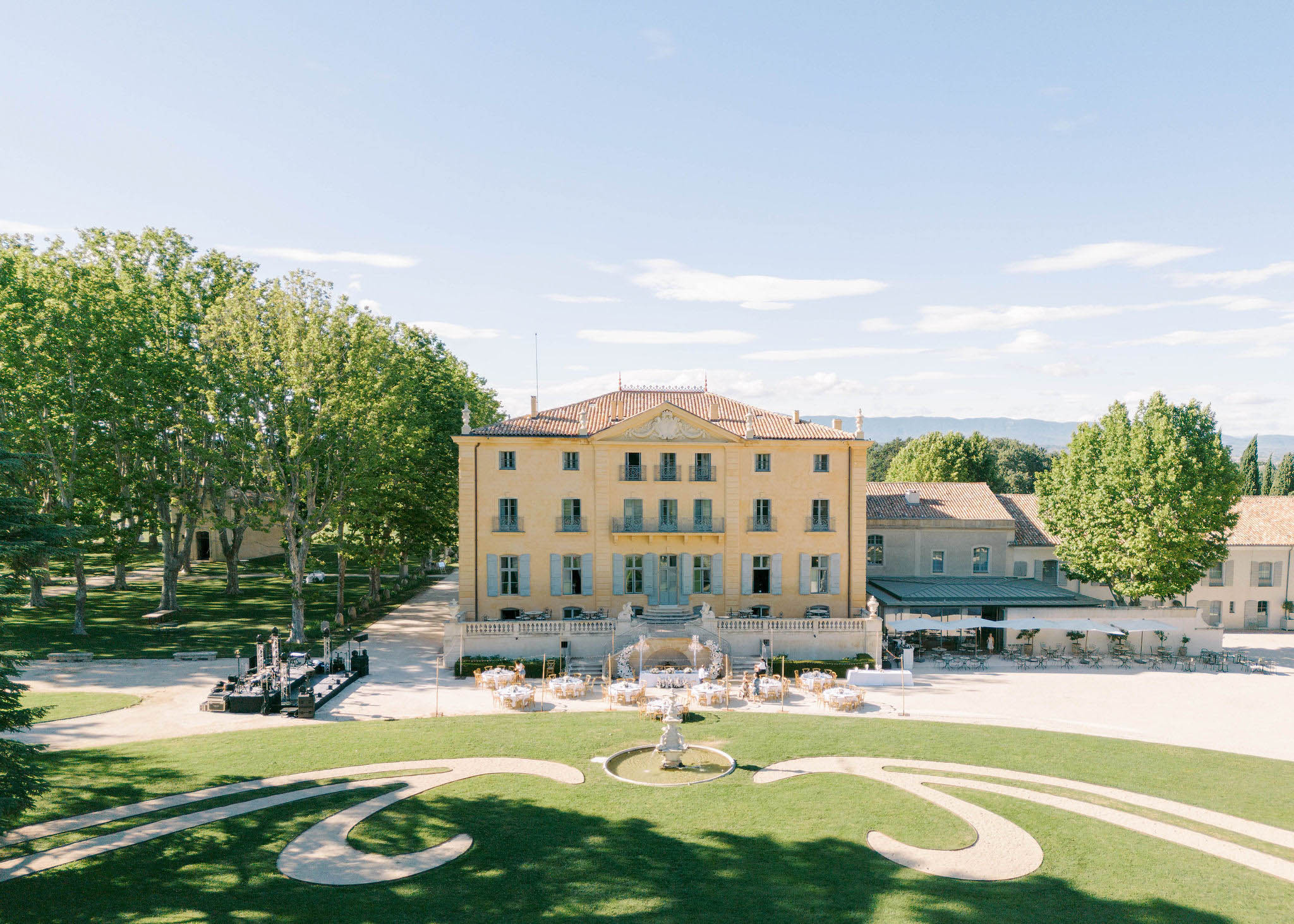 Aerial view of a yellow-ochre bastide chateau courtyard set with round reception tables around a stone fountain