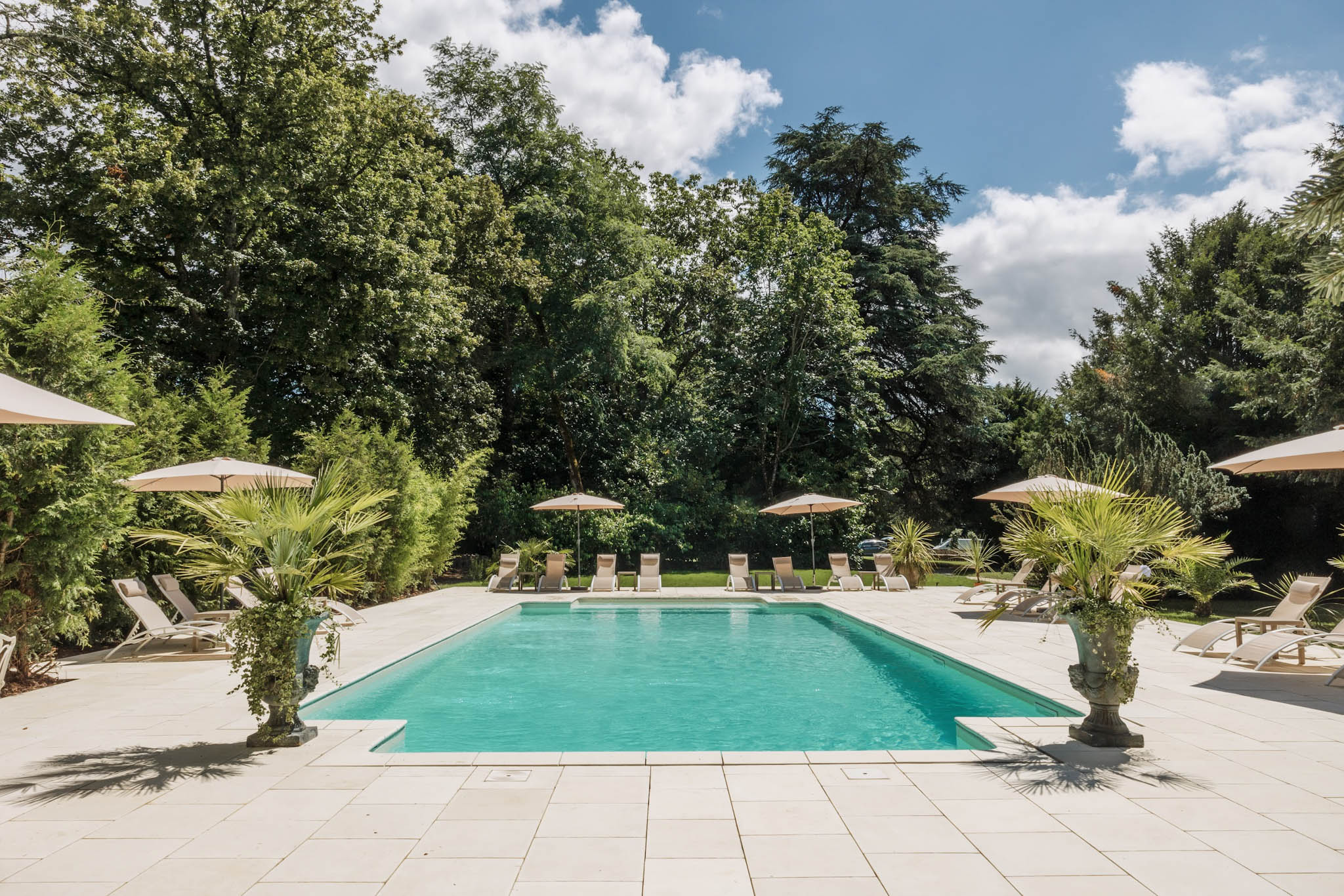 Rectangular pool with turquoise water surrounded by taupe sun loungers and beige parasols on a white stone terrace