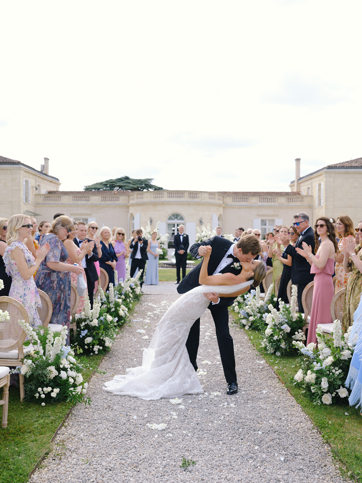 Groom dipping bride on gravel aisle with guests lining courtyard at classical cream stone venue