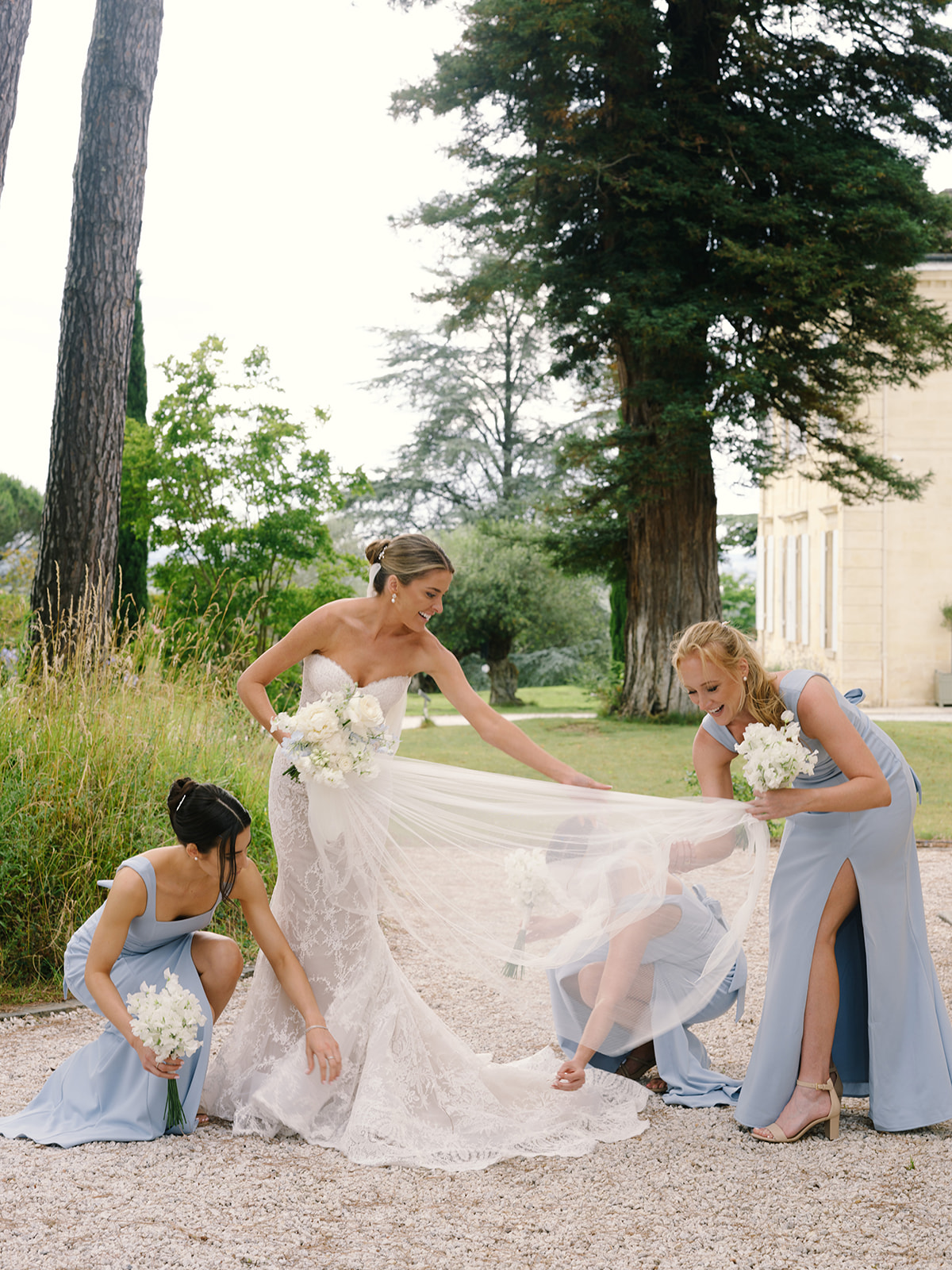 Bride in lace gown with three bridesmaids in periwinkle dresses arranging veil on tree-lined path
