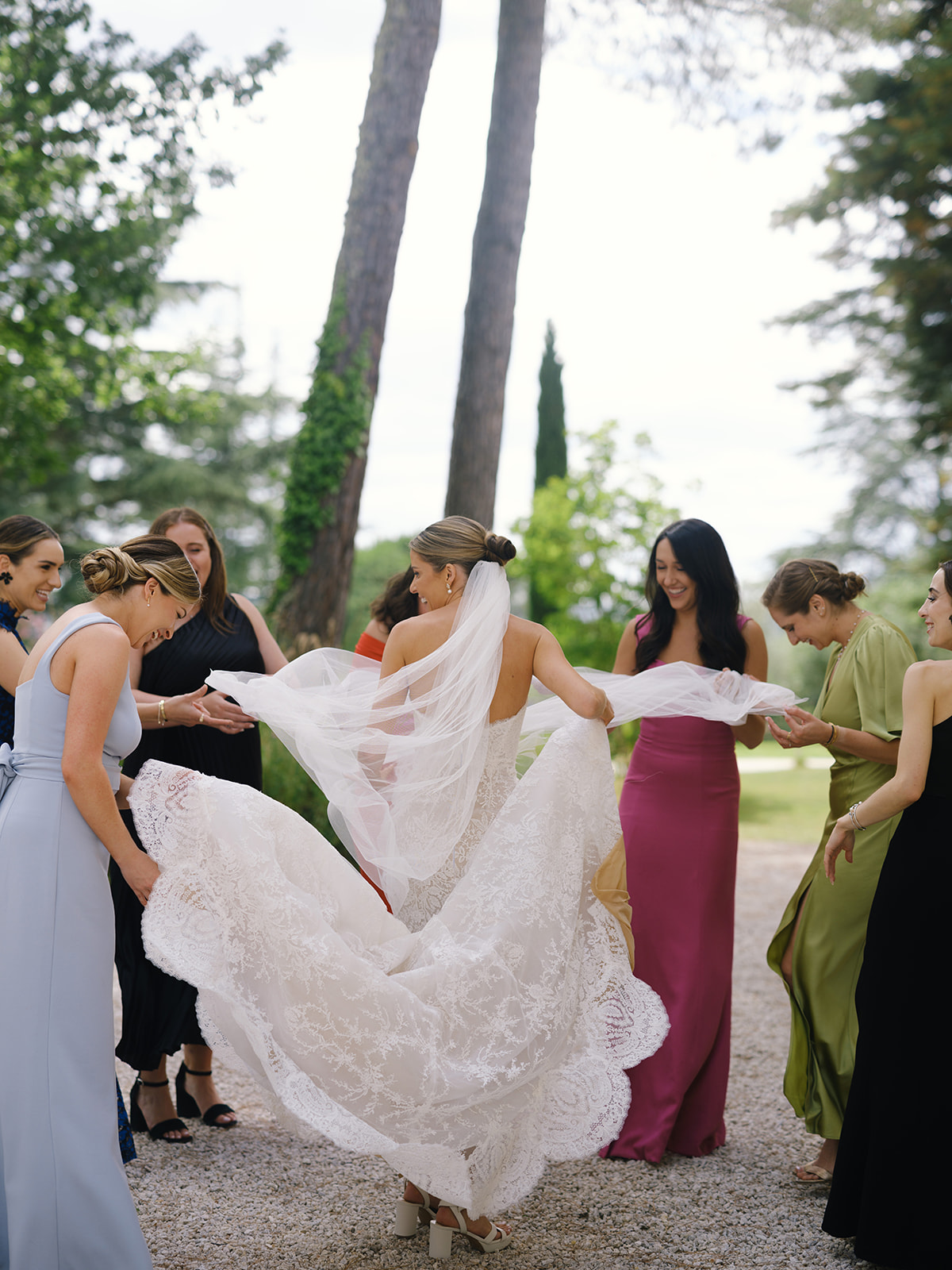 Bride with bridesmaids in mixed jewel-tone dresses displaying wedding train on cypress-lined garden pathway