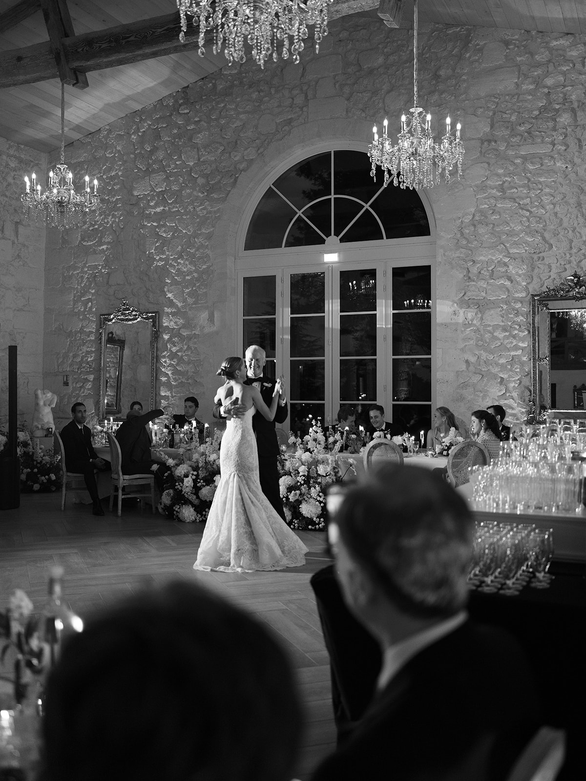 Black-and-white first dance in stone-walled venue with crystal chandeliers and arched windows