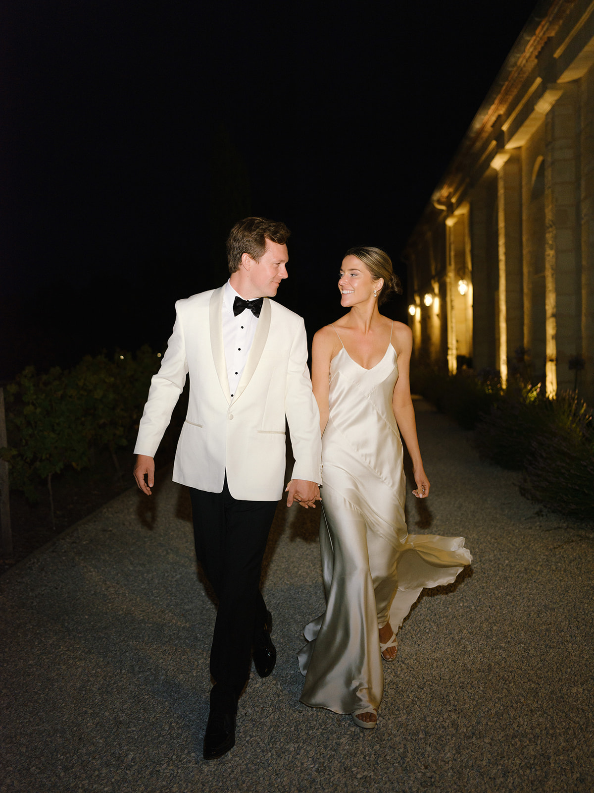 Bride and groom walking hand-in-hand along illuminated stone colonnade pathway at night after reception