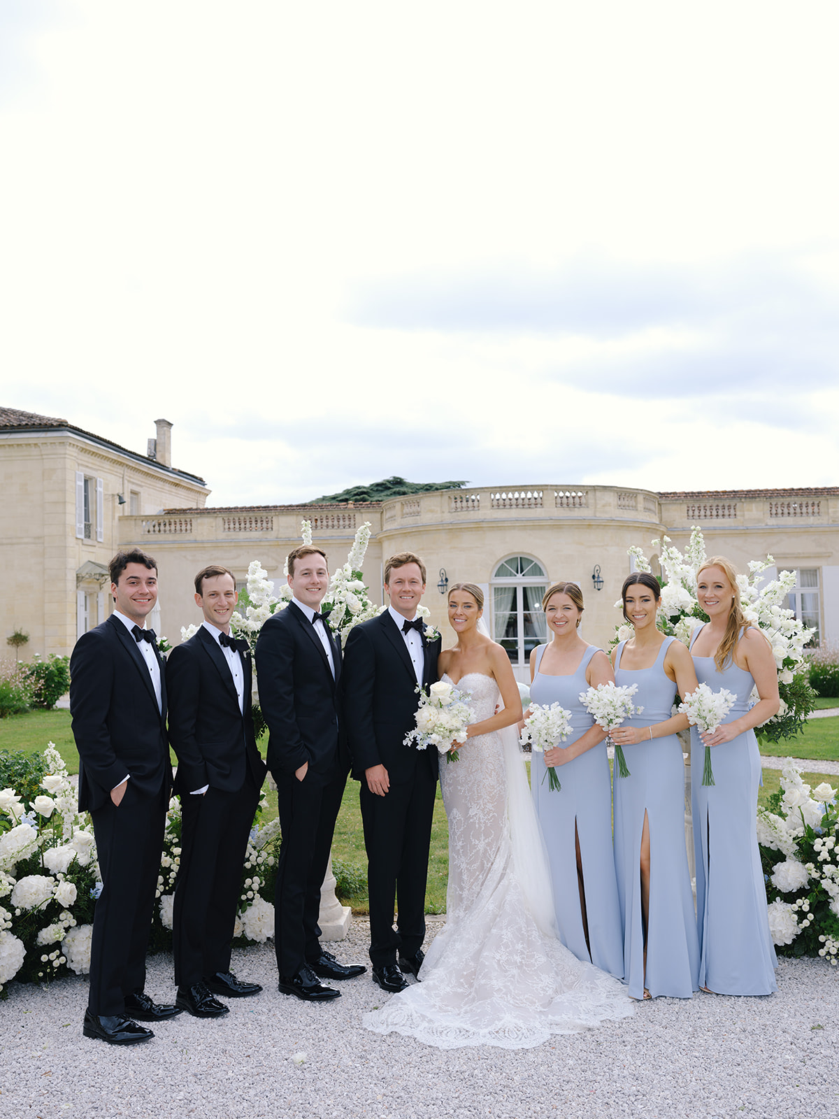 Bridal party portrait with bridesmaids in periwinkle gowns and groomsmen in black tuxedos before stone mansion