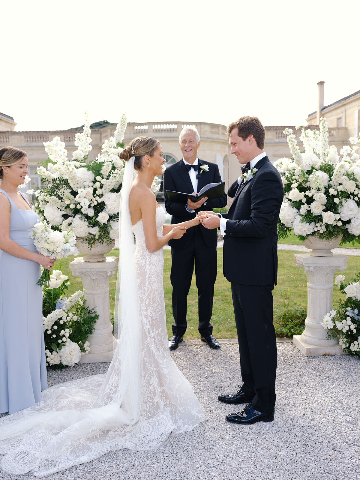 Bride and groom exchange rings before stone mansion, bridesmaid in lavender watches and white floral urns frame altar